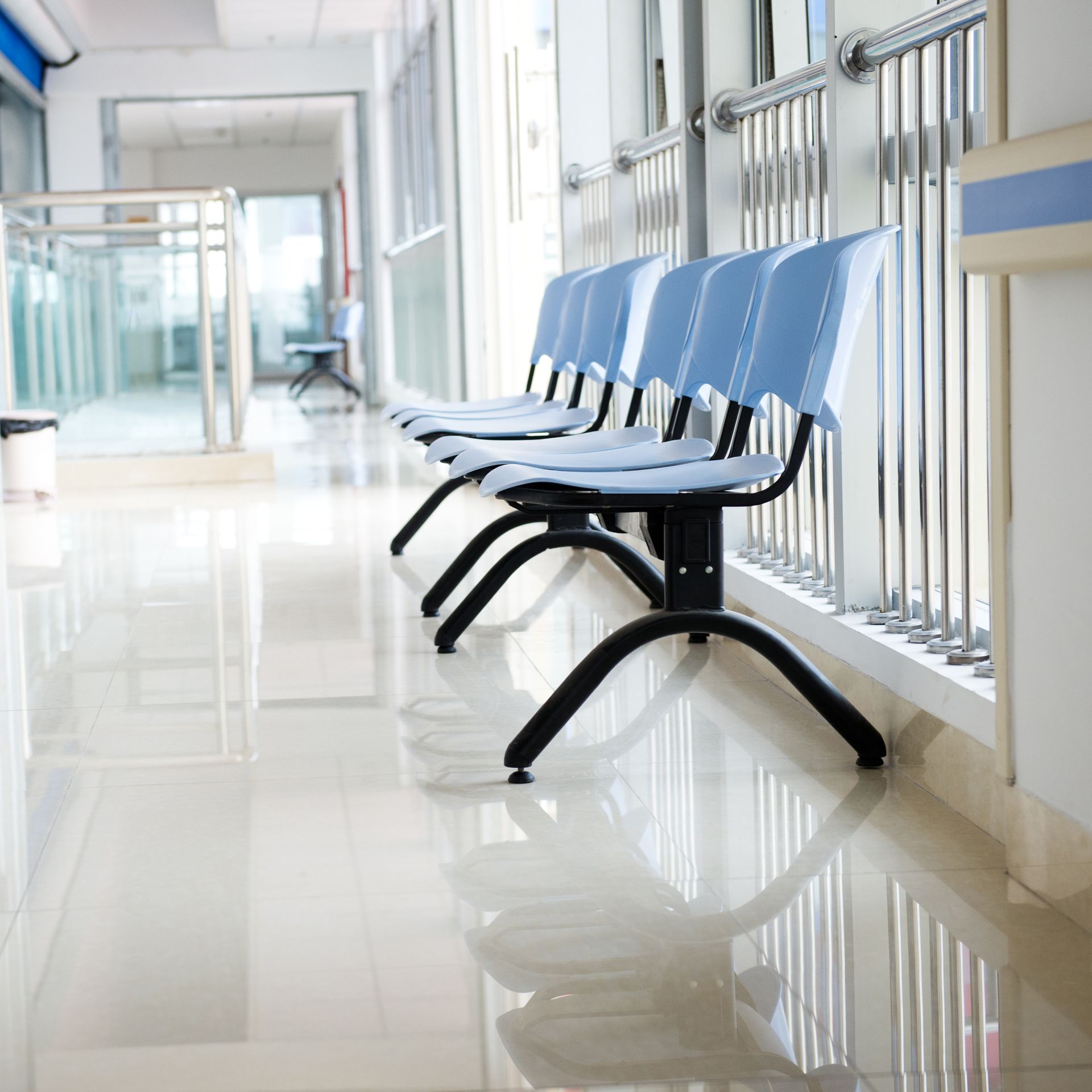 A row of light blue plastic waiting chairs with black metal legs positioned along a hallway in a medical facility.
