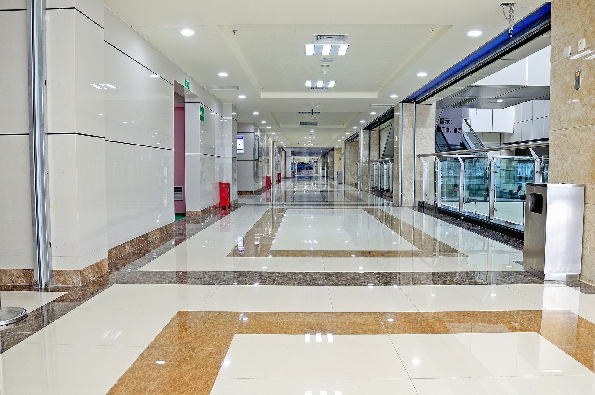 A brightly lit, empty corridor in a modern building with white tile floors, tan accent borders, and glass railings.