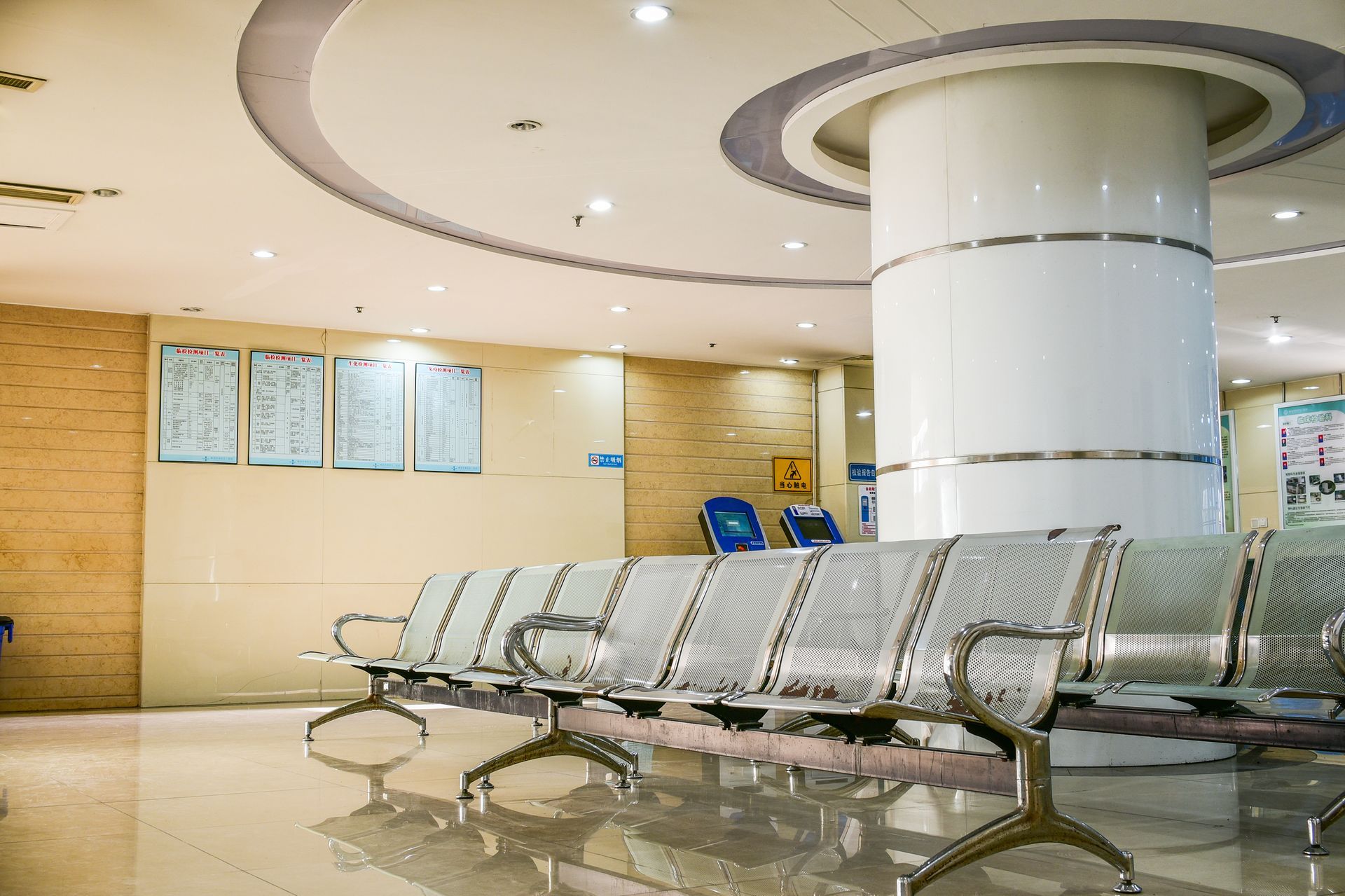 A brightly lit, modern hospital waiting area with rows of empty silver metal chairs and a large white circular column.