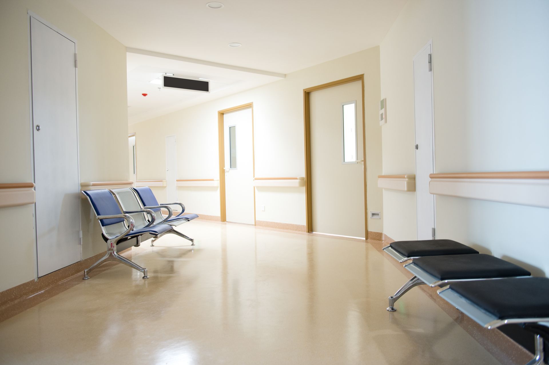 A sterile hospital corridor with cream-colored walls, polished floors, and rows of empty waiting chairs.
