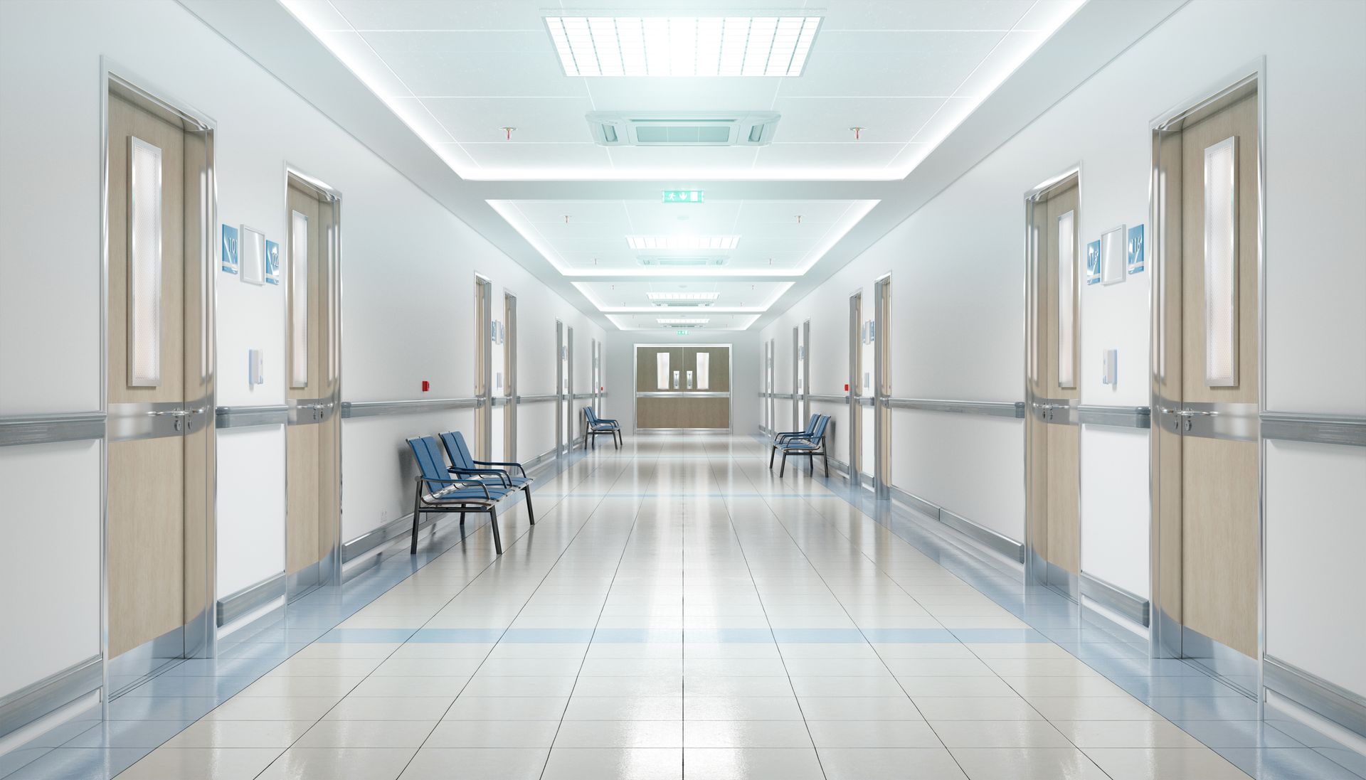 A long, bright, modern hospital corridor with white walls, tiled floors, and several blue chairs lining the sides.
