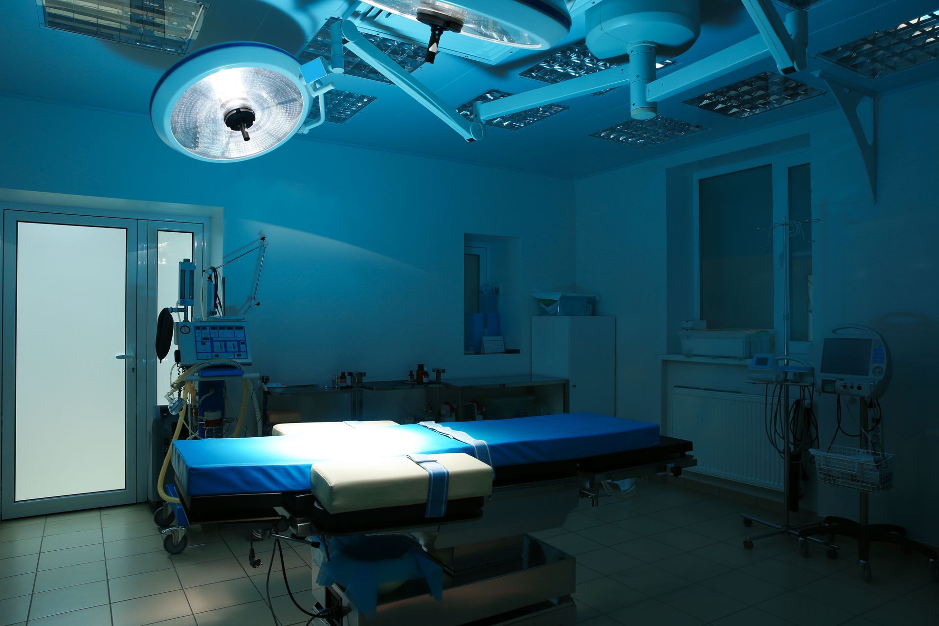 An empty surgical suite with a blue-draped operating table, medical equipment, and large overhead surgical lights.