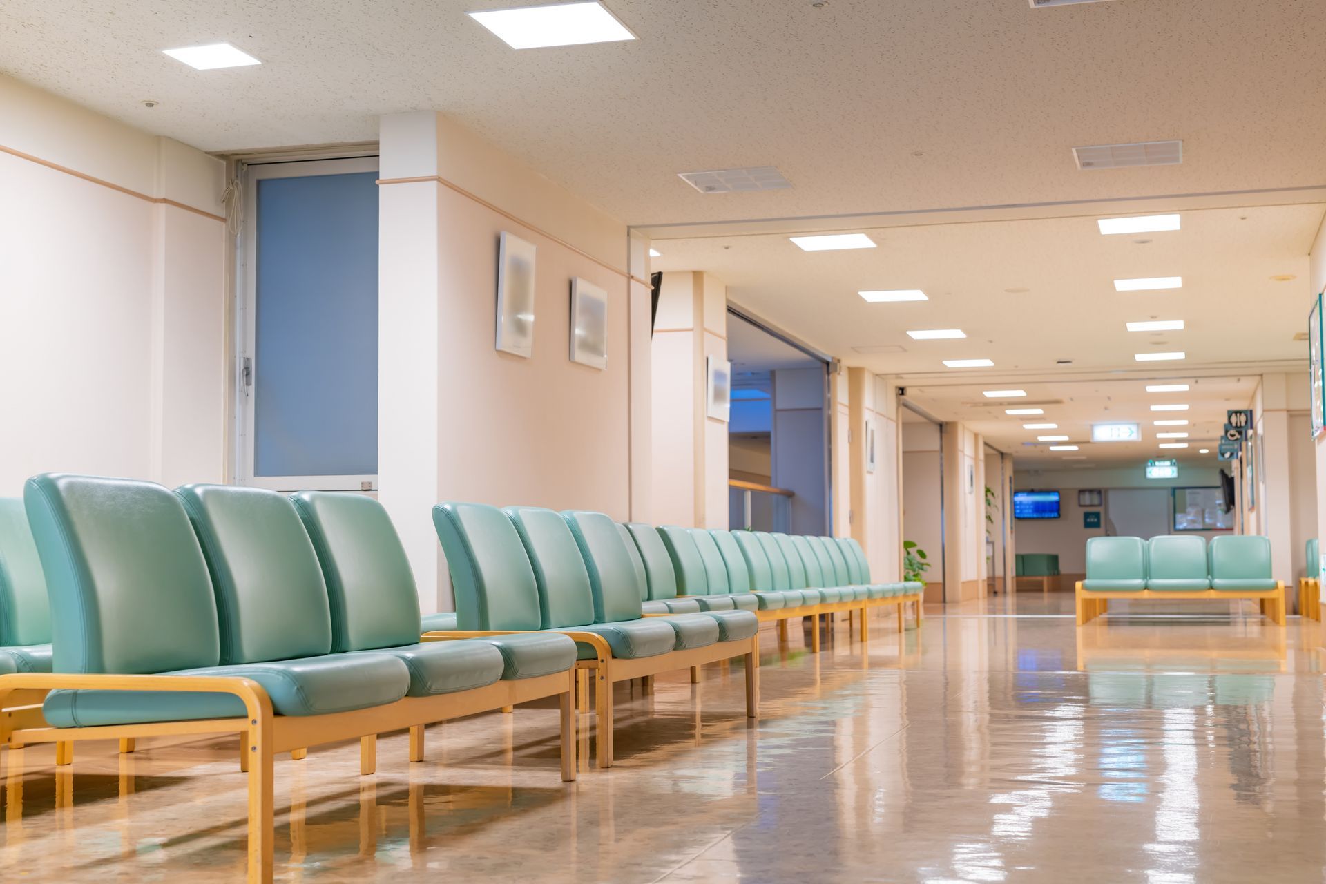 A quiet, empty hospital waiting room with rows of light teal chairs on a polished floor under bright ceiling lights.