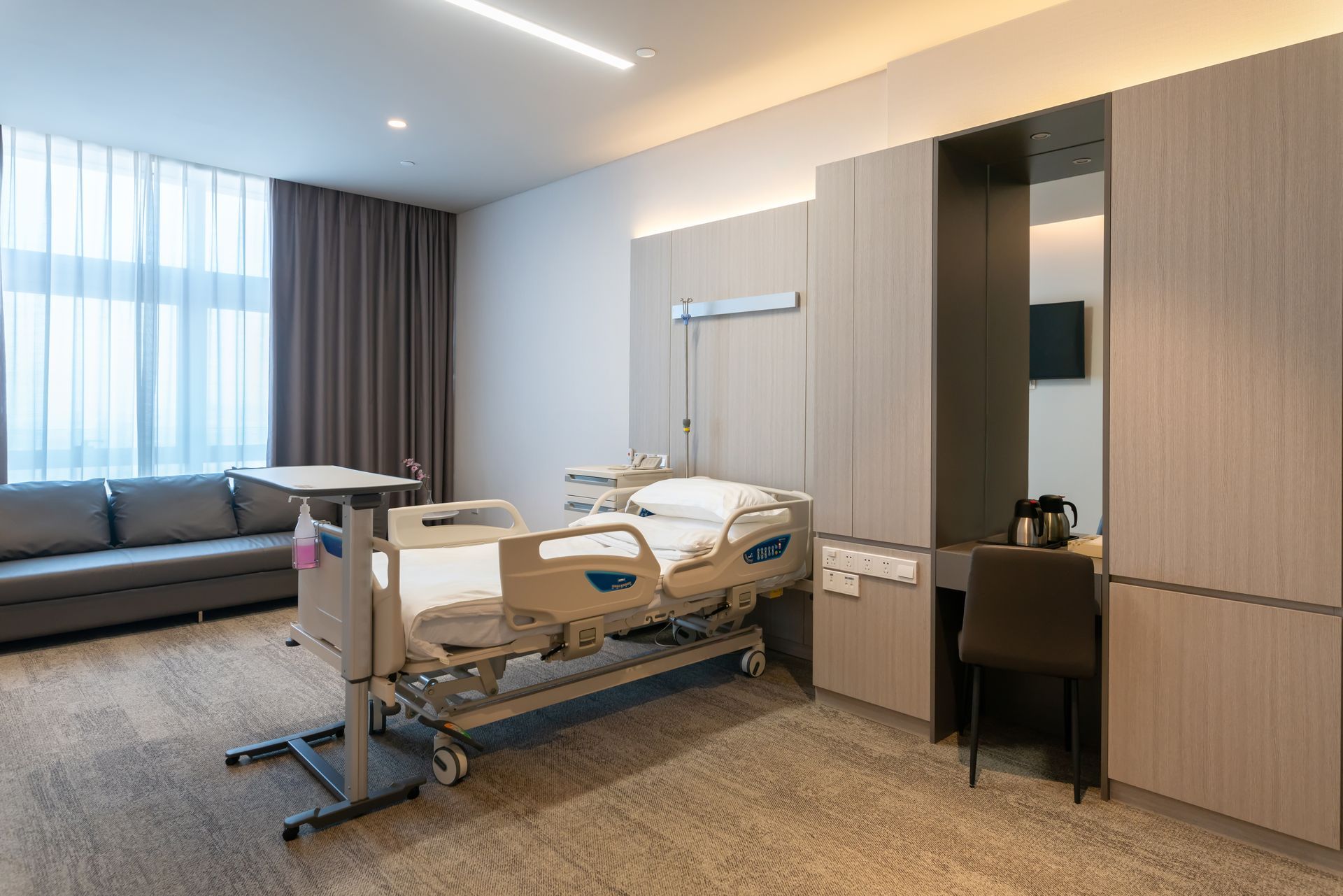 A modern, neutral-toned hospital room with a patient bed, a sofa for visitors, and a built-in desk and wardrobe unit.