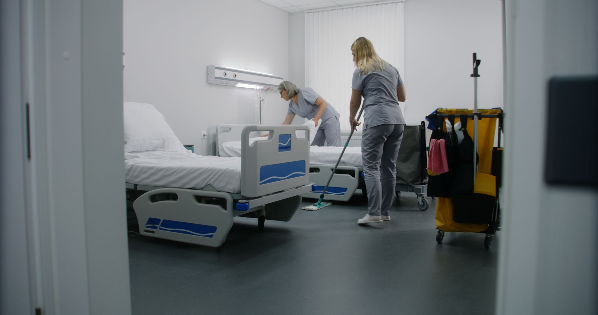Two hospital staff members in gray uniforms clean a hospital room with beds and a yellow supply cart.