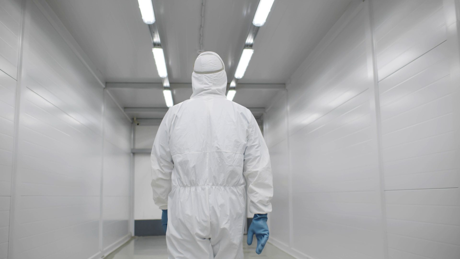 A person in a white protective suit walks down a brightly lit, sterile white corridor.