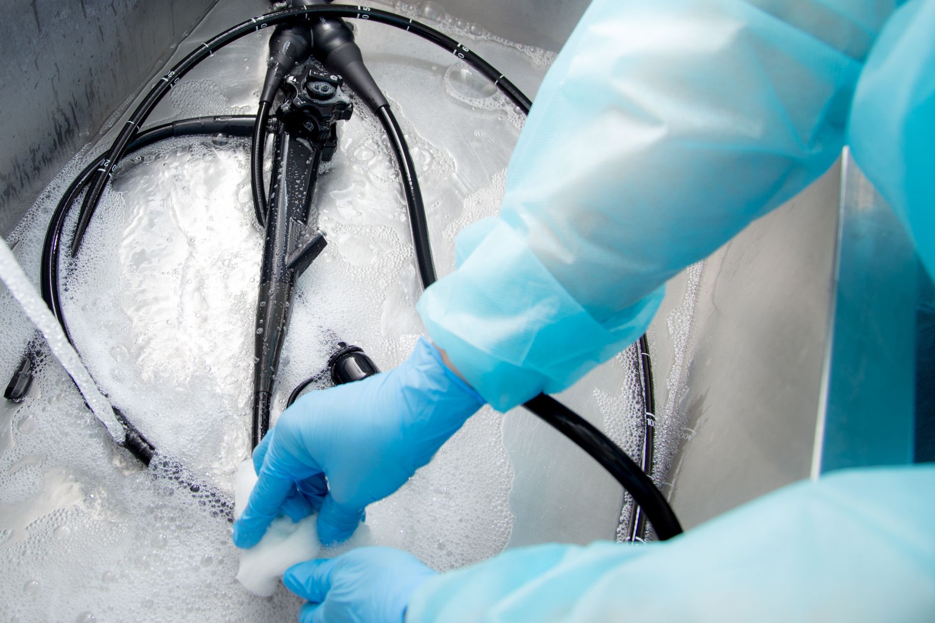 A healthcare worker wearing blue gloves and protective gear manually cleaning an endoscope in a sink with soapy water.