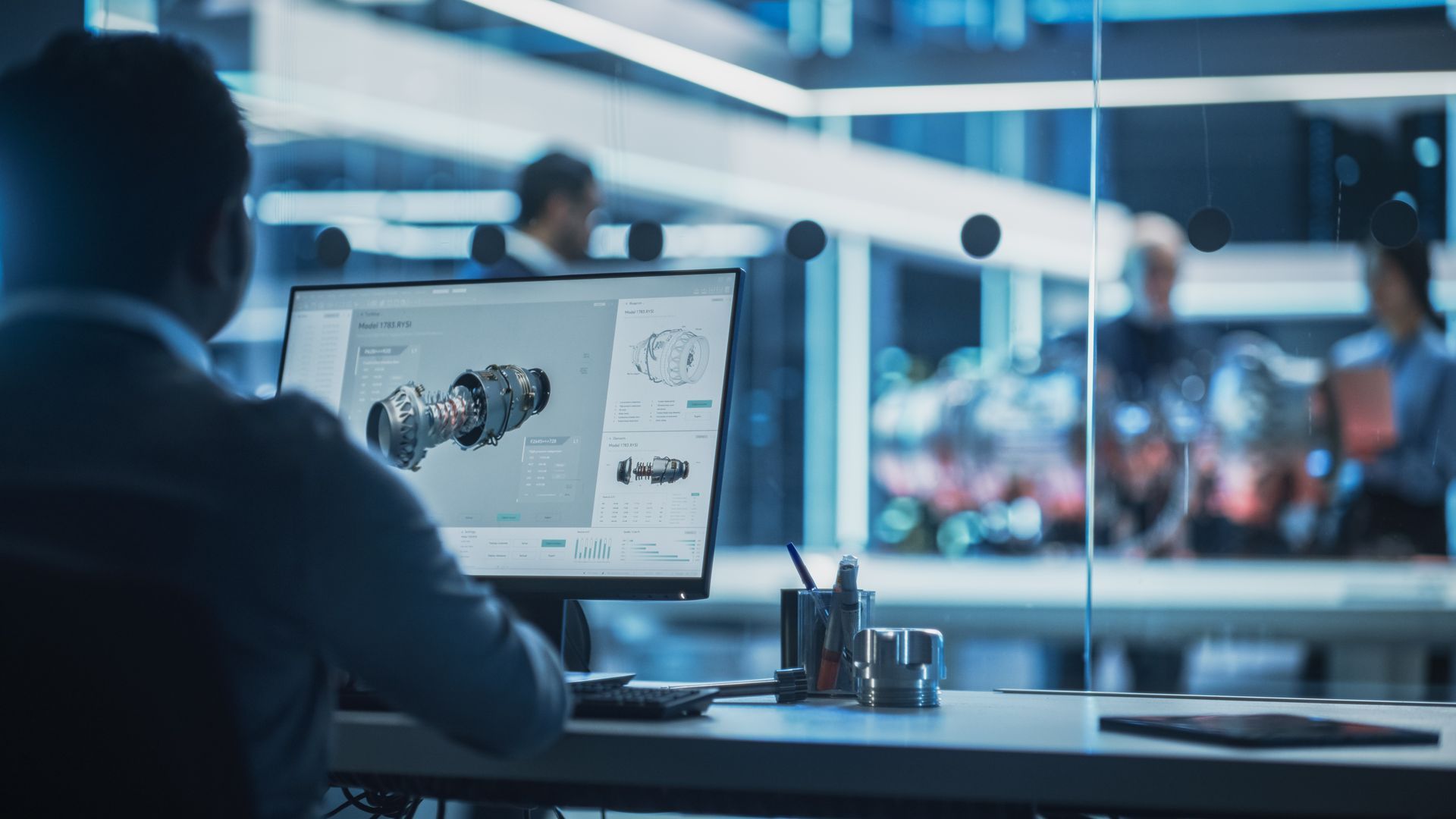 A person at a desk in a modern office monitors 3D mechanical designs on a computer screen, with colleagues visible nearby.