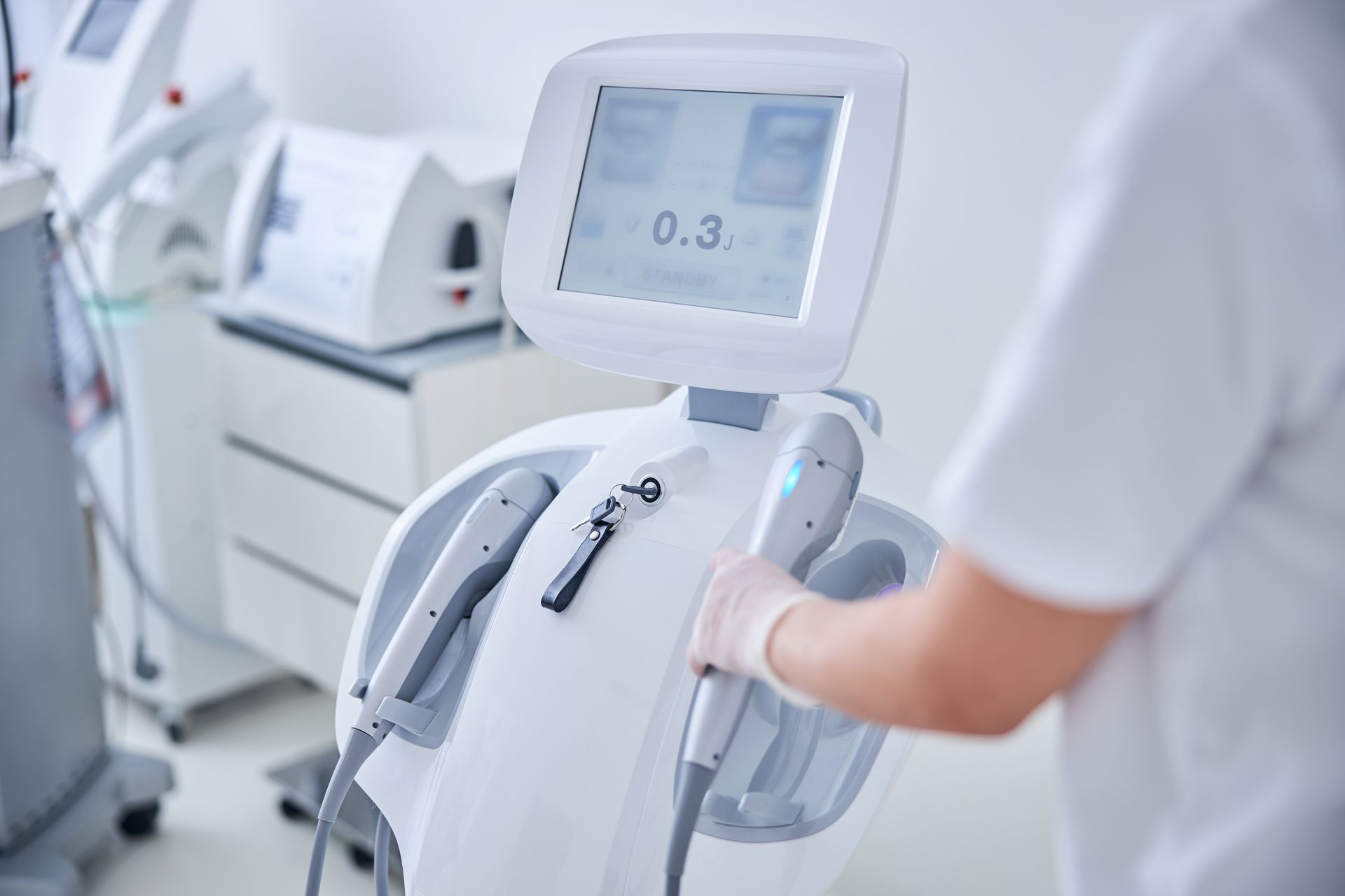 A person in a white uniform prepares a modern medical aesthetic device with a digital display in a clinic setting.