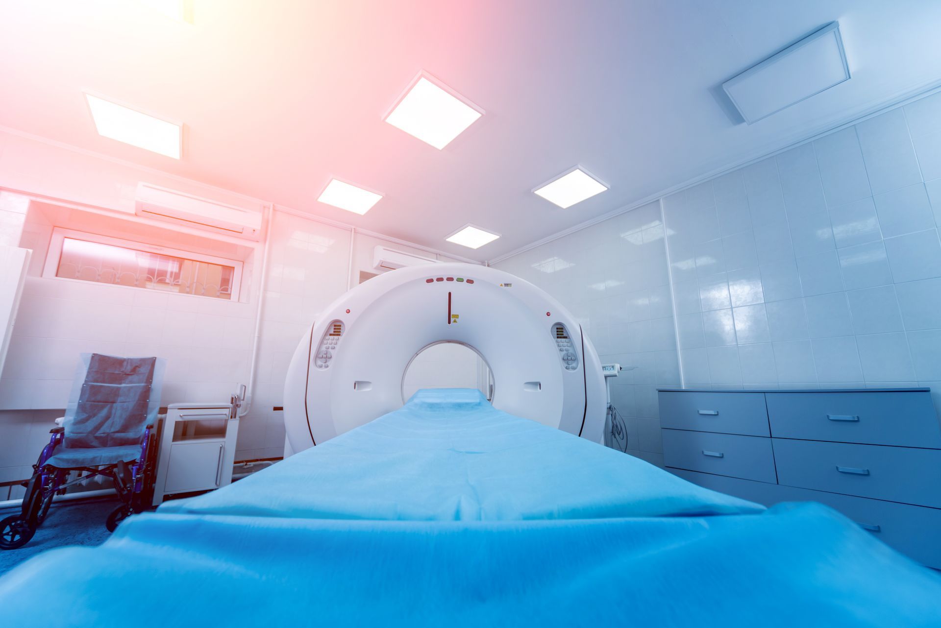 A medical CT scan machine in a sterile clinical room with a light blue examination table and a wheelchair in the corner.