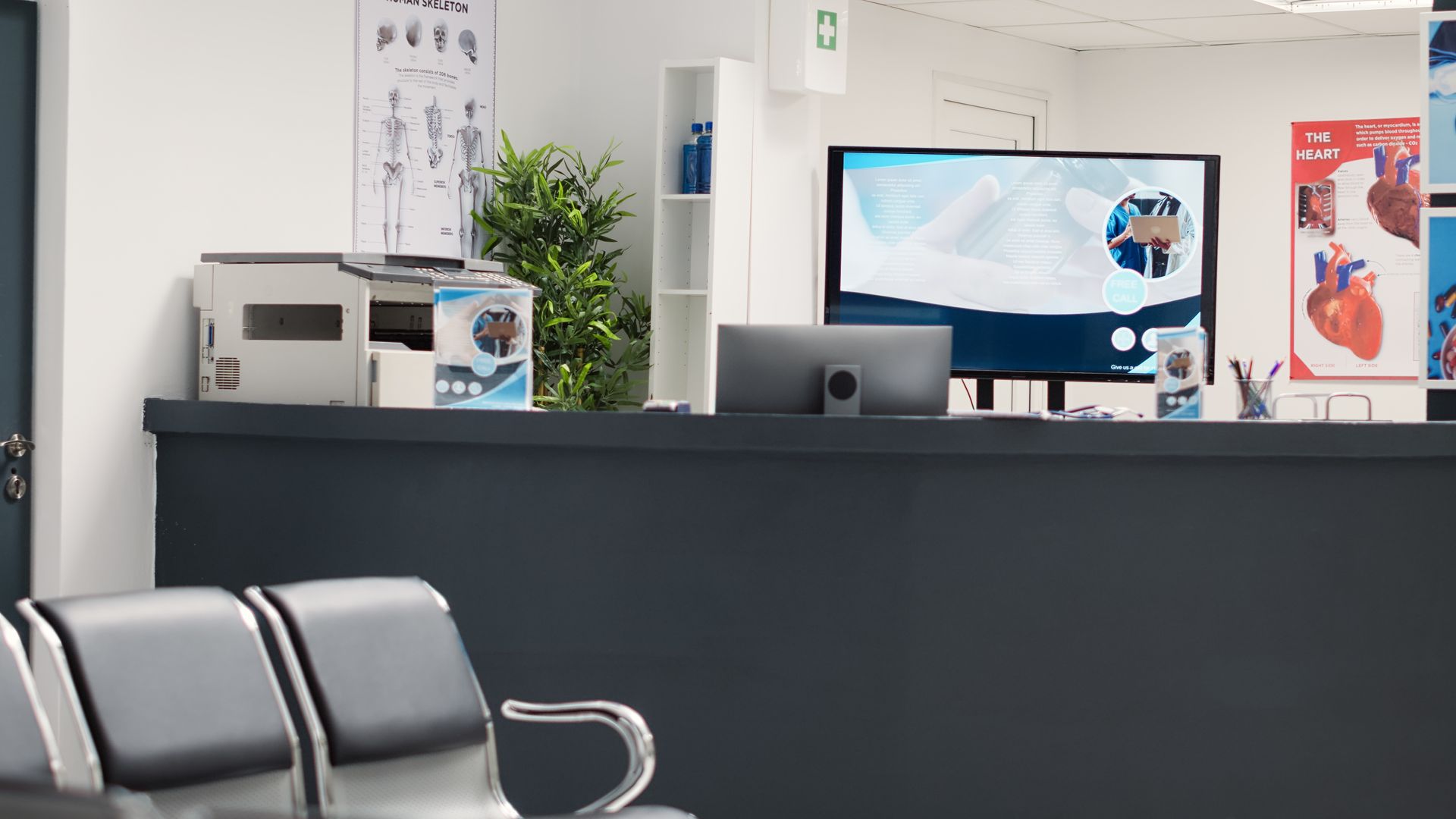 Reception desk with a computer and printer in a medical office, featuring anatomical charts and a waiting area.