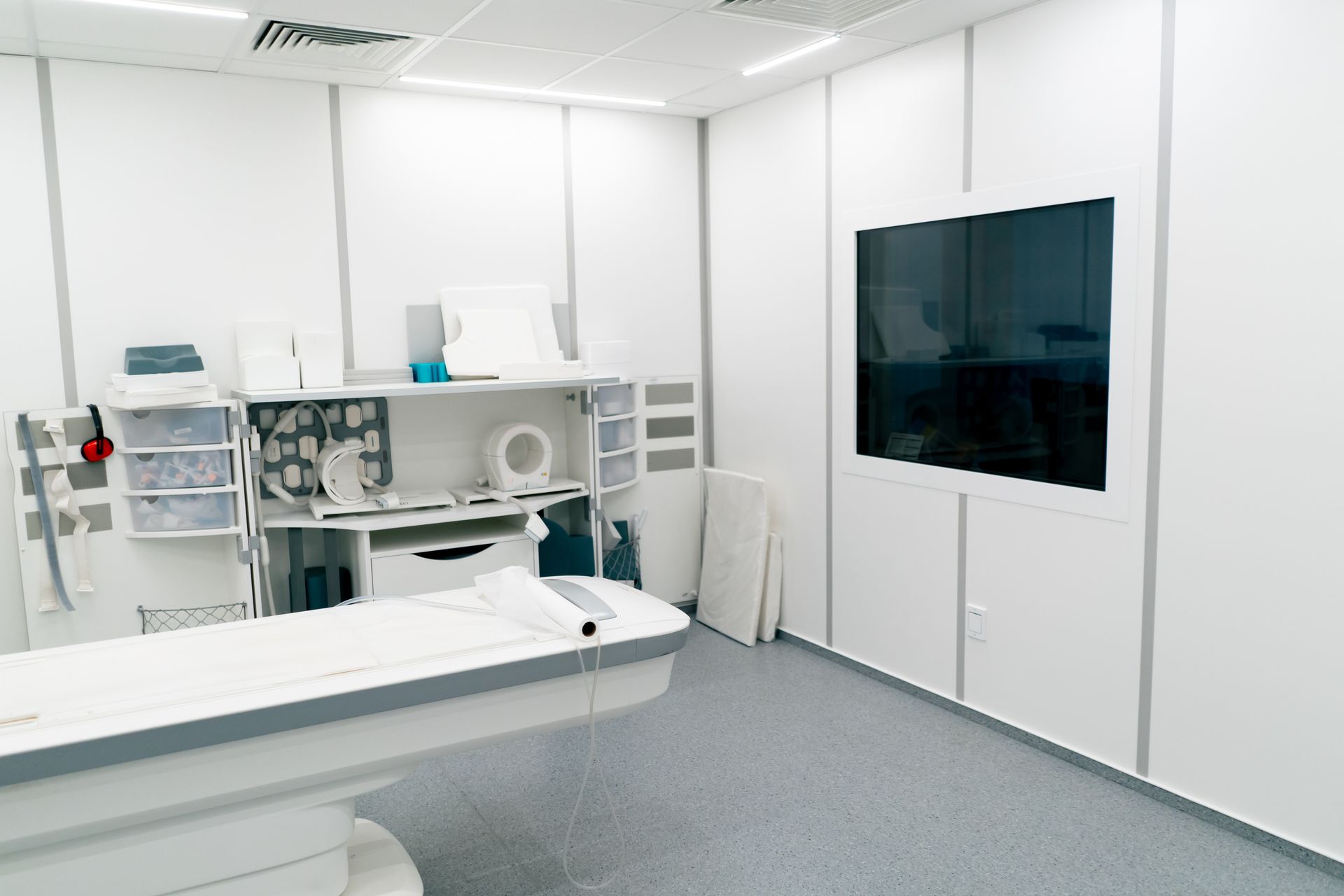 A bright, minimalist medical examination room with a white exam table, storage cabinets, and a dark wall-mounted monitor.