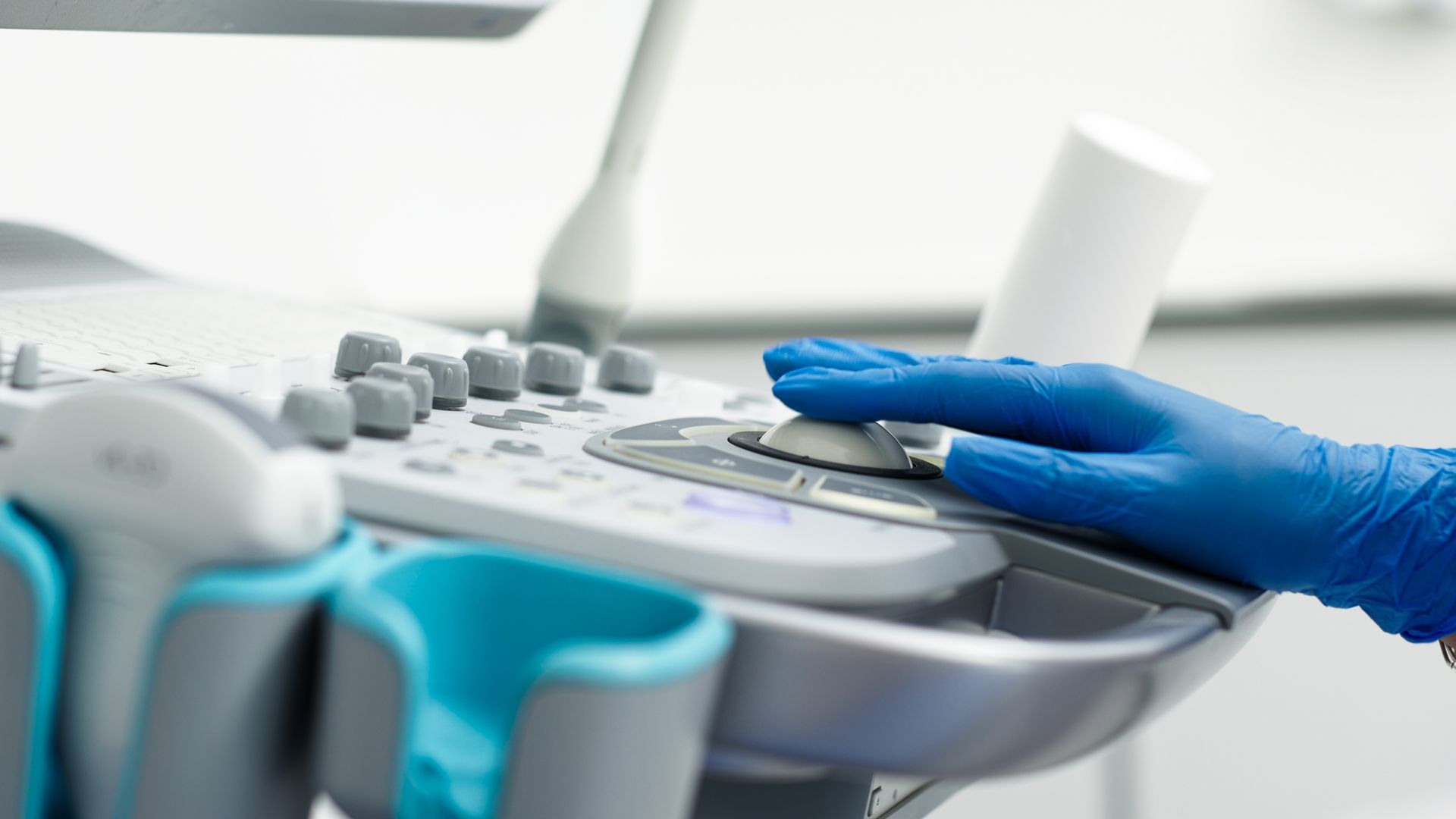 A hand wearing a blue medical glove adjusts the control dial on an ultrasound machine console.