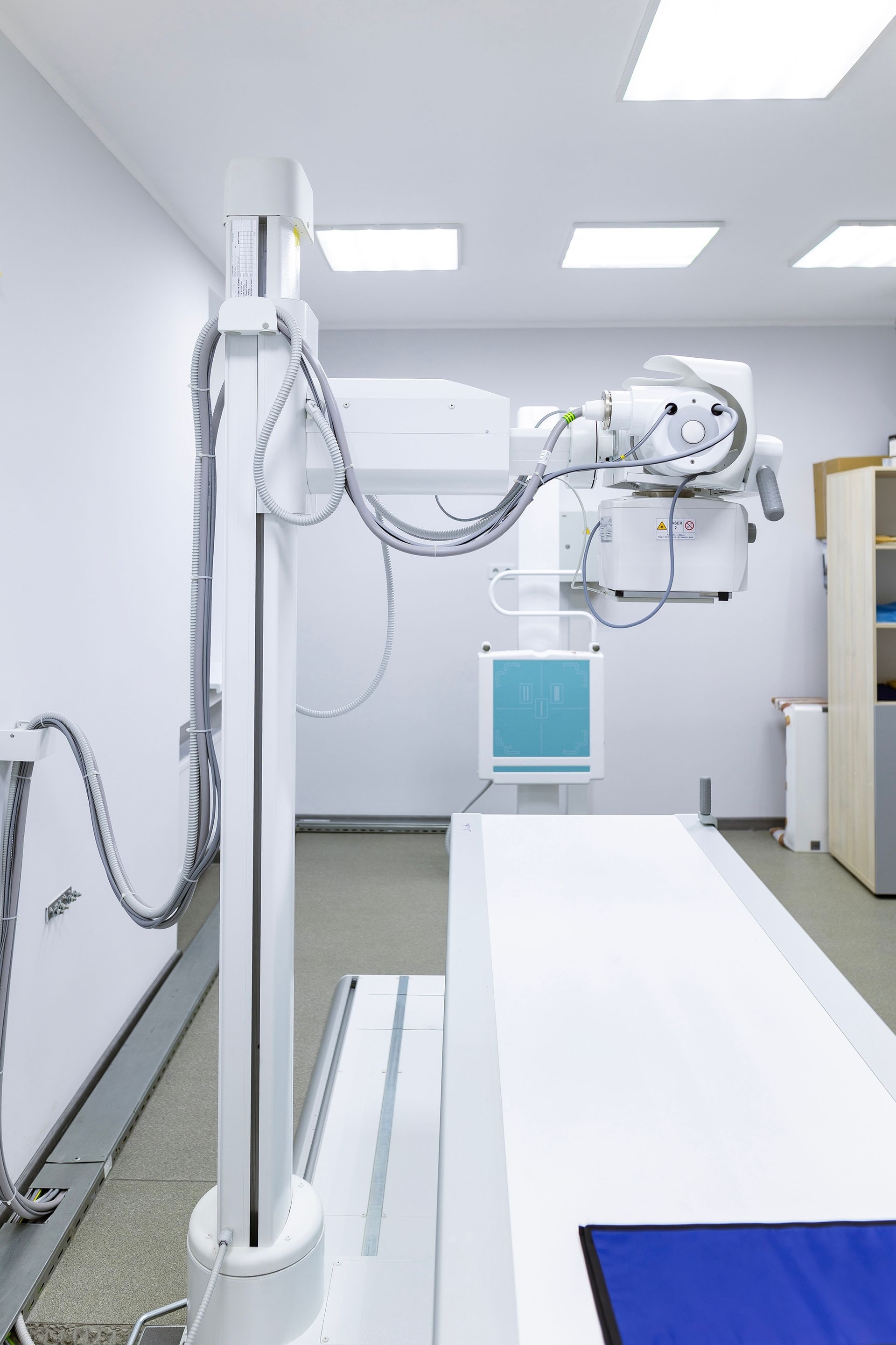 A digital X-ray machine with a white overhead tube and table inside a clinical, brightly lit examination room.
