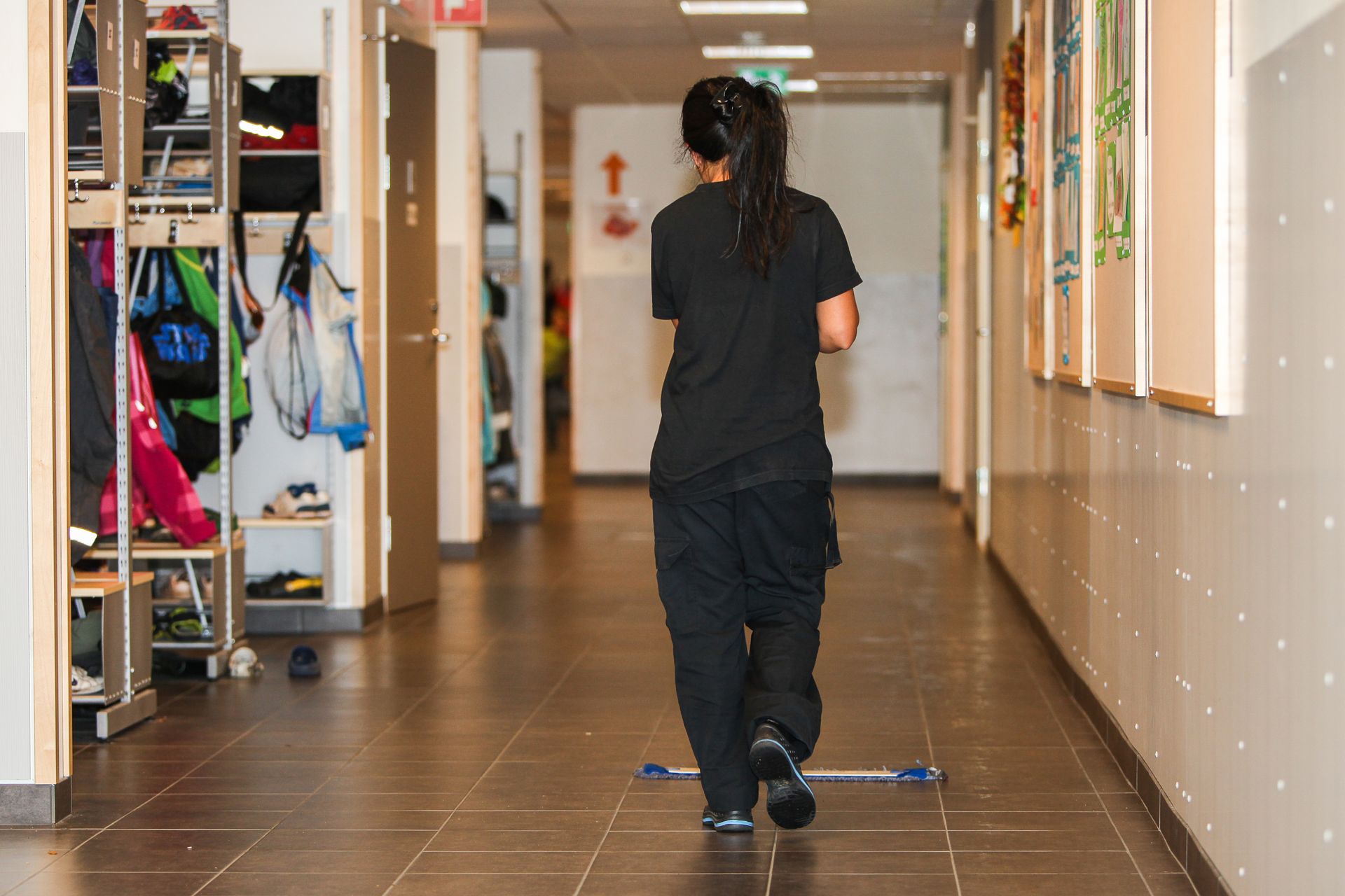 A person in all-black clothing walks down a school hallway past storage cubbies and art on the walls.