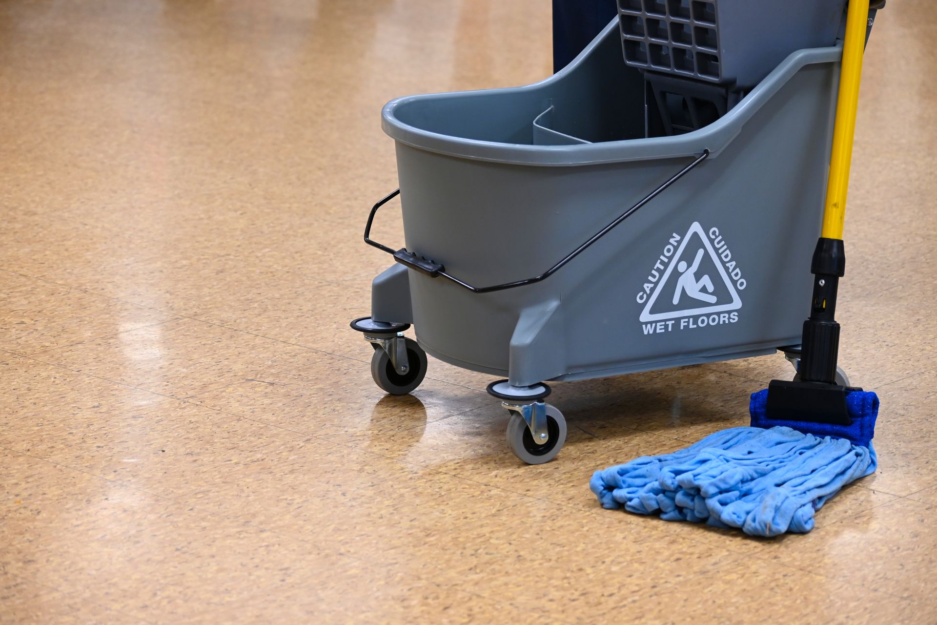A grey mop bucket with a warning sign sits on a wet floor next to a blue mop on a yellow handle.
