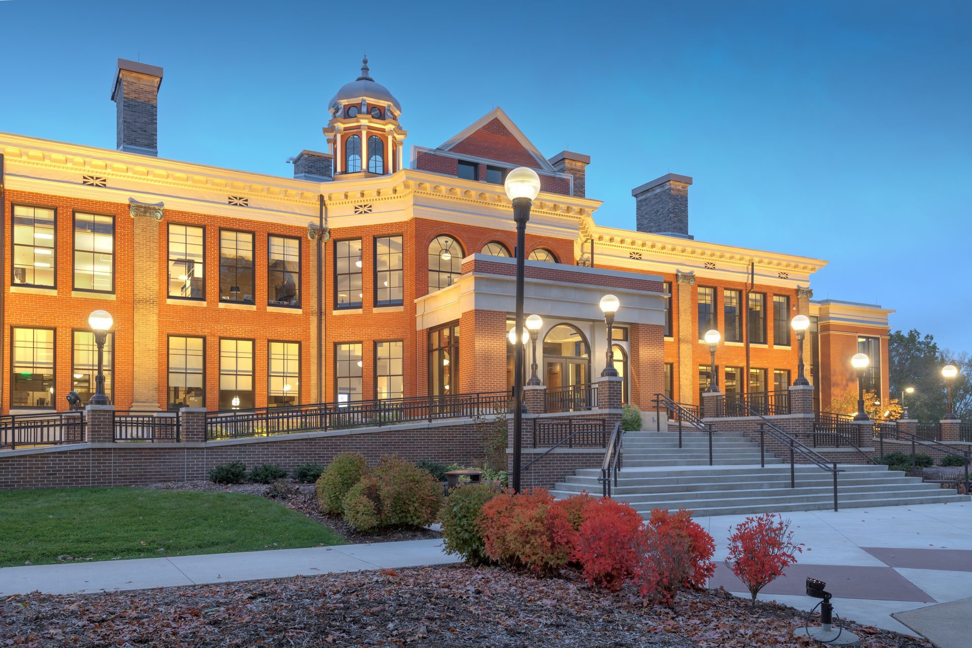 Illuminated brick building with a central cupola and stairs, surrounded by landscaped bushes at dusk.