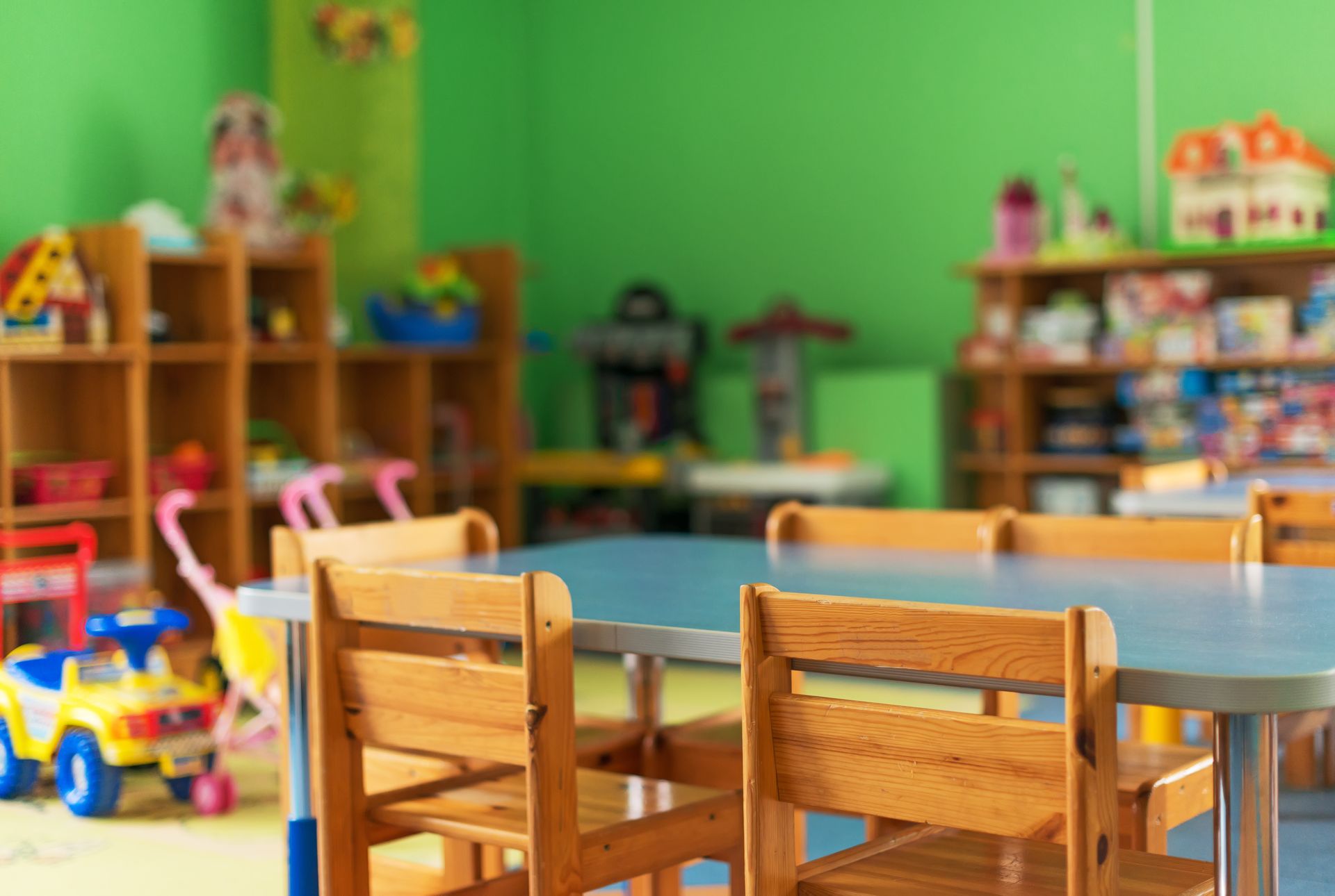 A brightly lit classroom with a table, wooden chairs, shelves full of toys, and green walls.