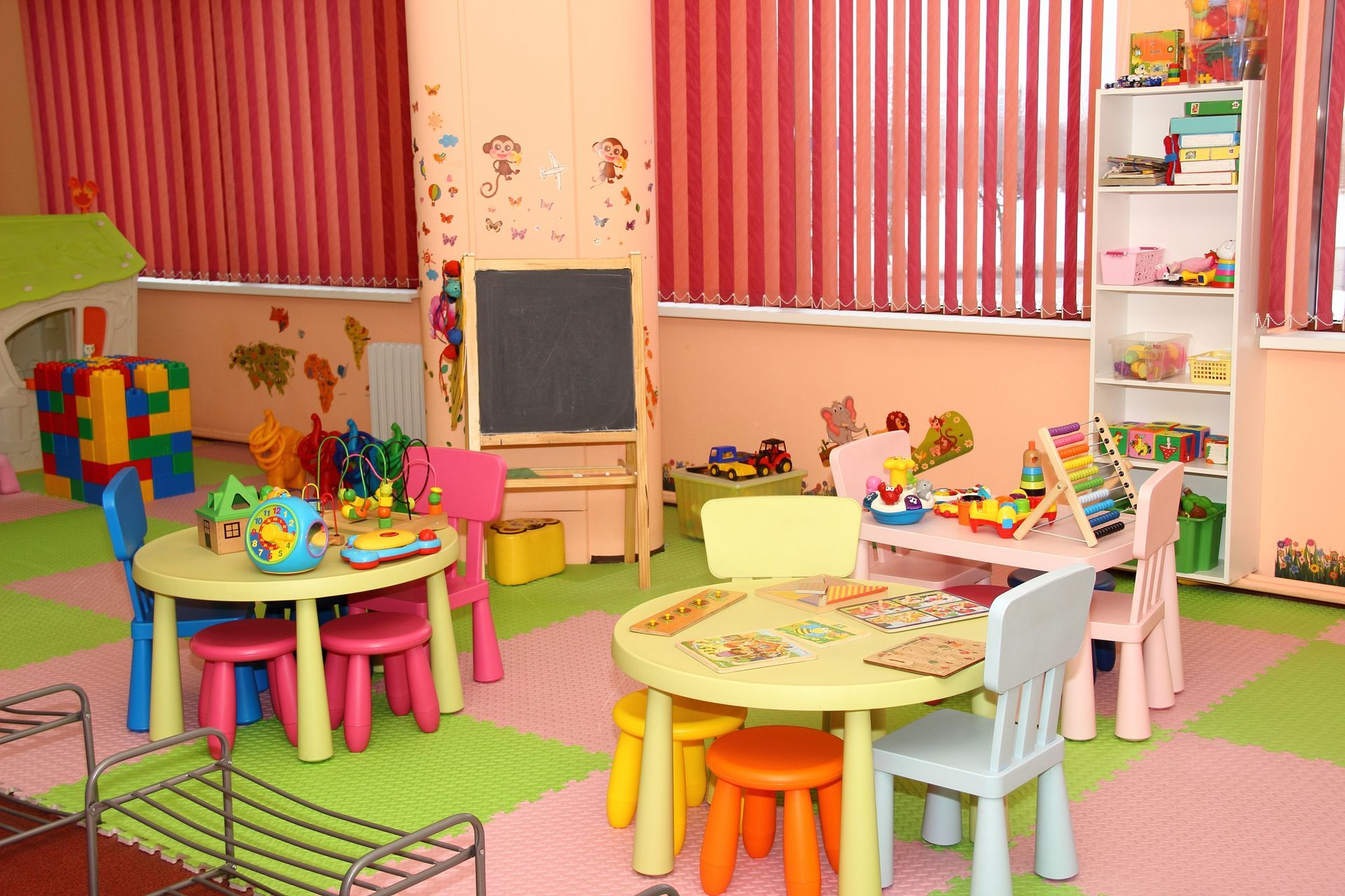 A bright playroom featuring two yellow tables with colorful chairs, a small chalkboard, and shelves filled with toys.