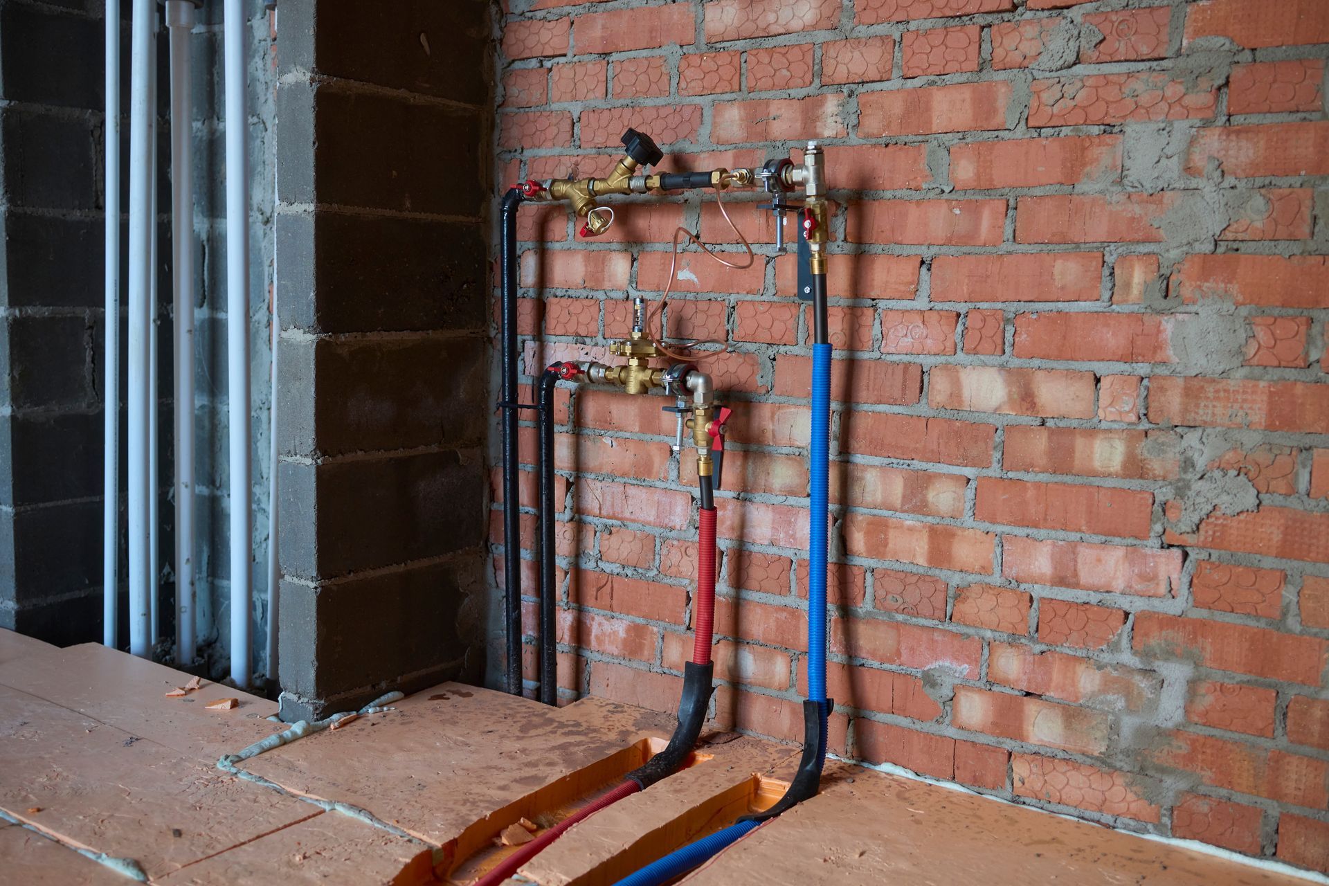 A plumbing manifold setup with brass valves and red and blue insulated pipes attached to an unfinished brick wall.
