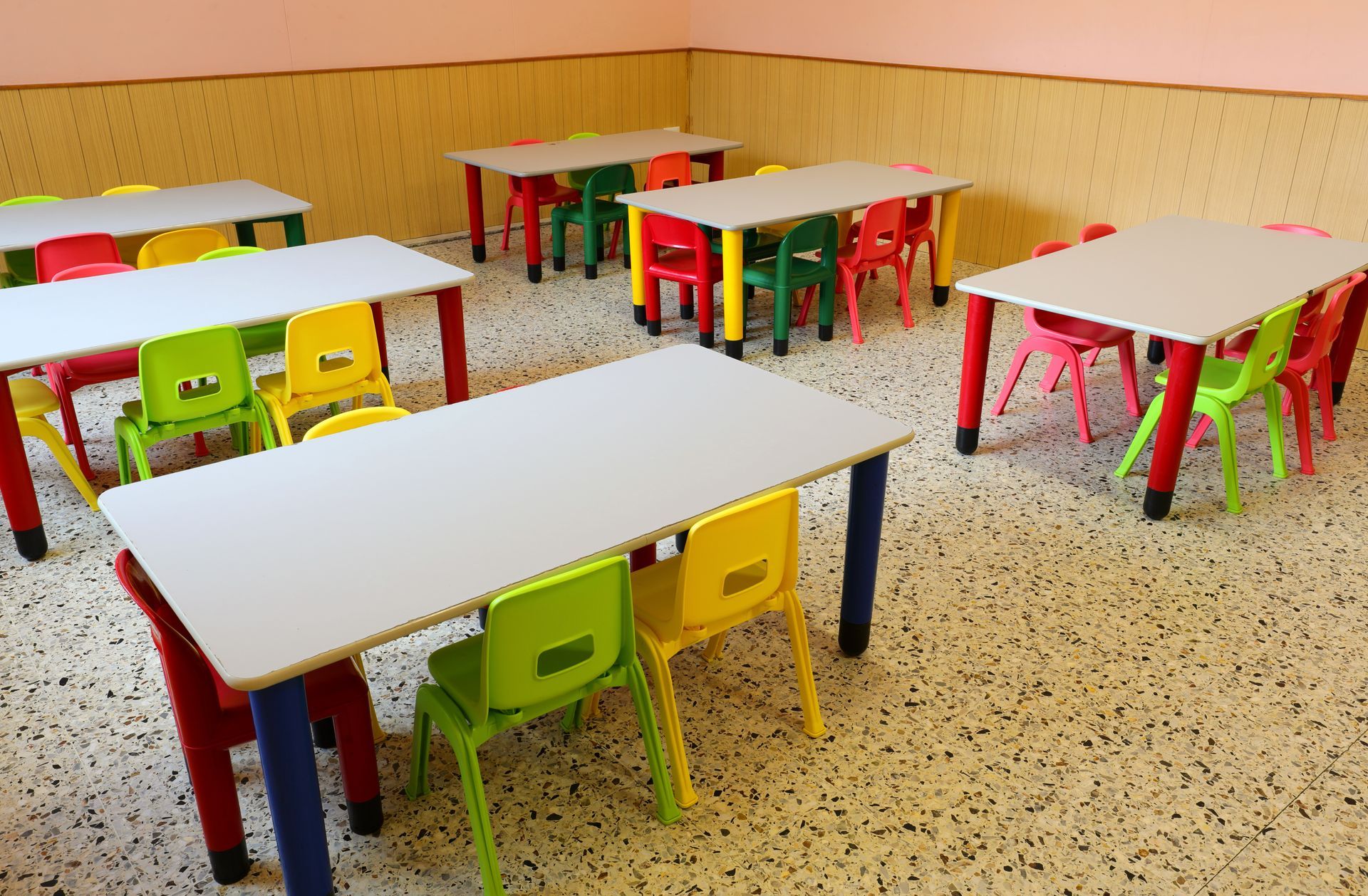 A classroom with several white tables and colorful chairs on a speckled floor.