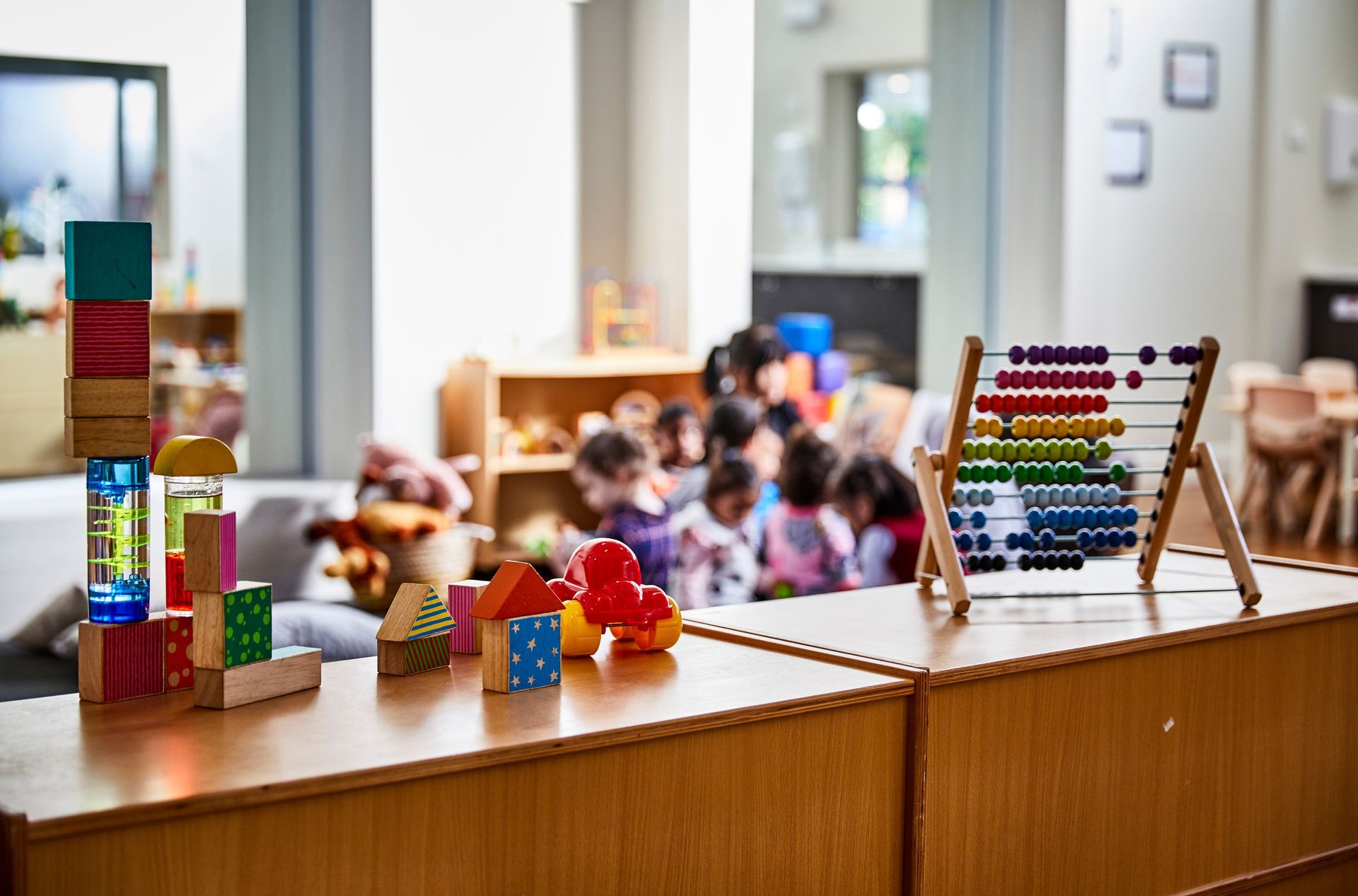 Colorful building blocks and a wooden abacus sit on a shelf in a brightly lit classroom with children in the background.
