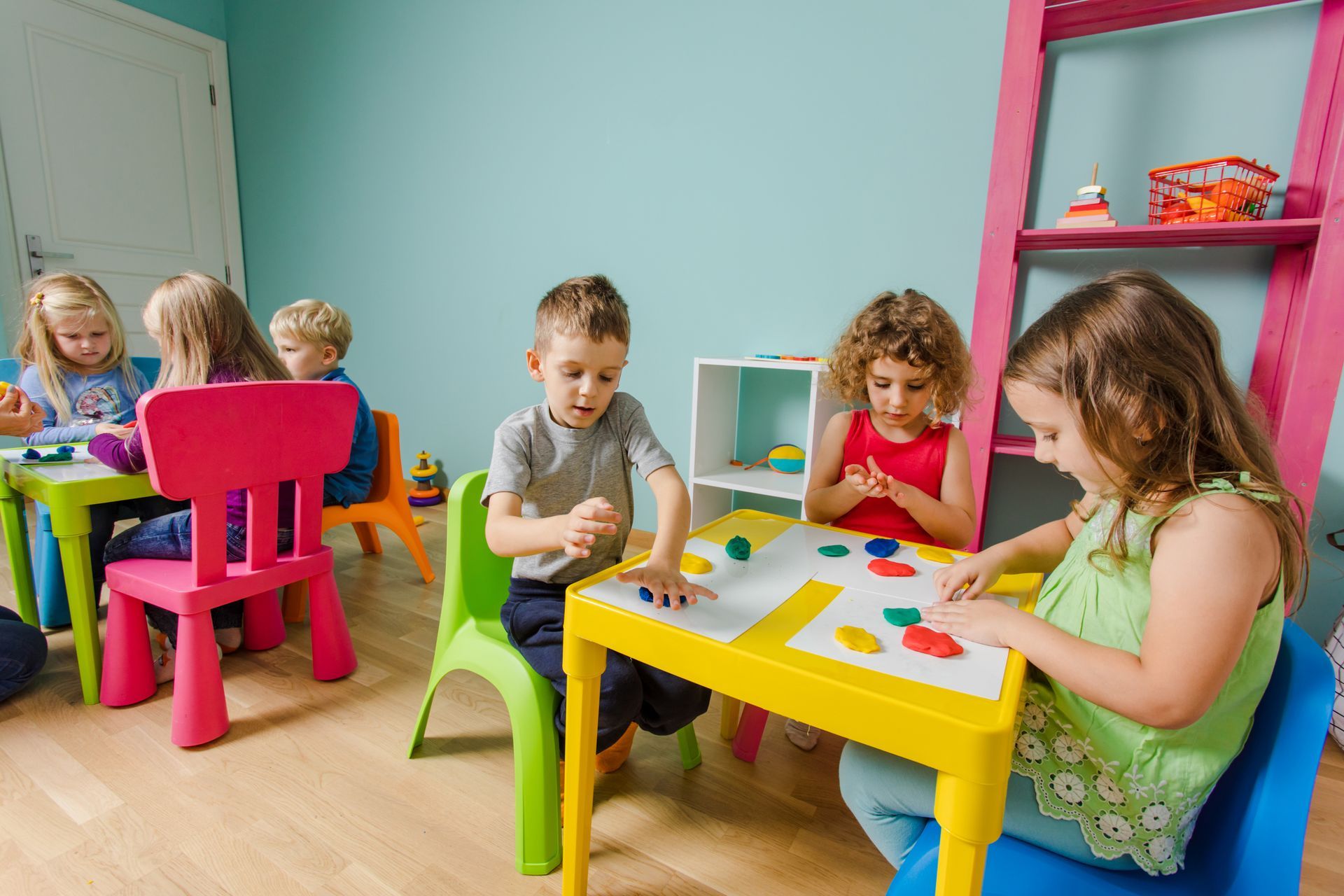 Children sitting at small, brightly colored tables in a classroom, engaged in play with modeling clay.