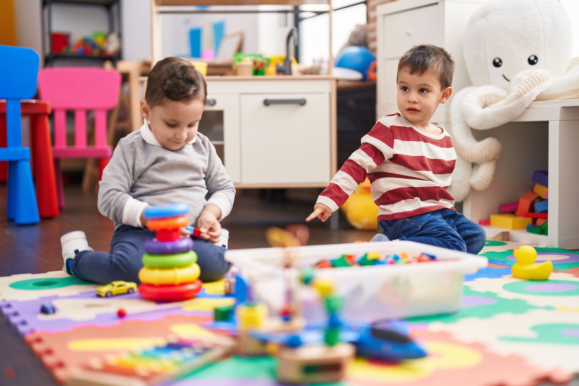 Two children sitting on a colorful floor mat in a playroom, one stacking colorful rings and the other pointing at toys.