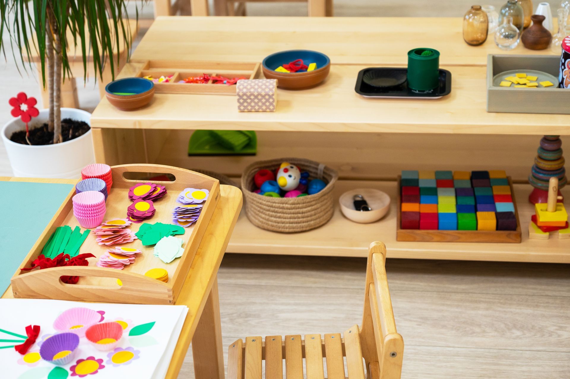 A wooden shelf filled with educational toys, Montessori materials, and a craft project on a child's desk in a classroom.