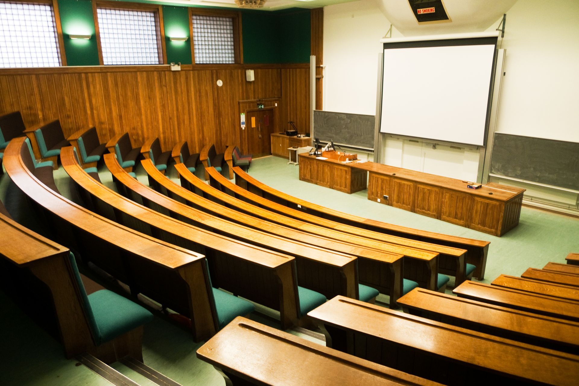 A lecture hall with curved wooden rows of seats facing a teacher’s desk, chalkboard, and a large projection screen.
