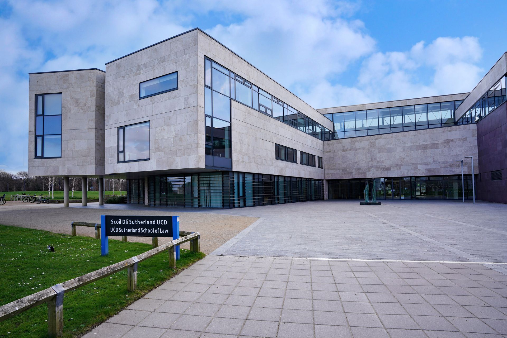 A modern, light-colored stone building with large glass windows, situated next to a paved courtyard and a grassy lawn.