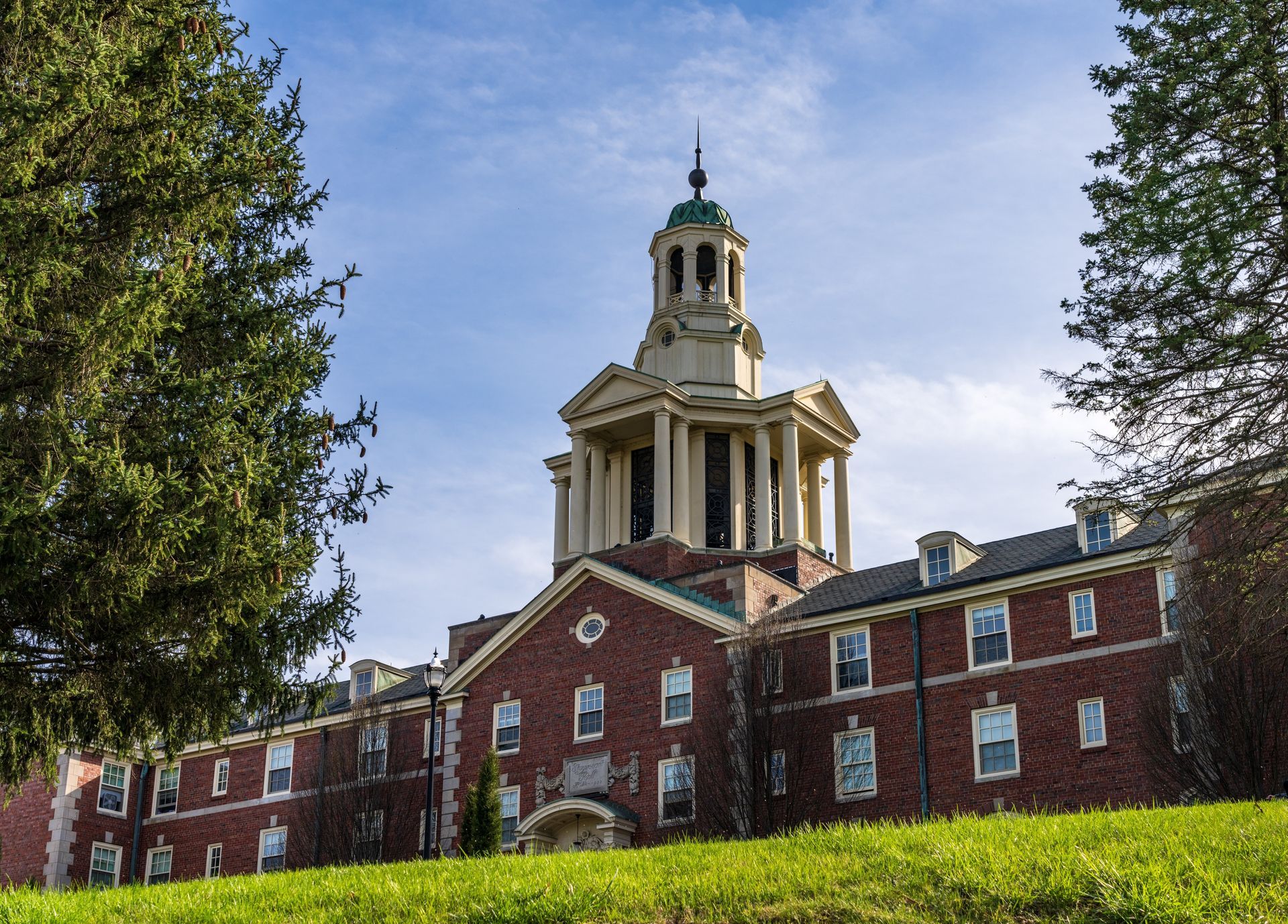 A brick academic building with a prominent central bell tower, framed by trees under a bright blue sky.