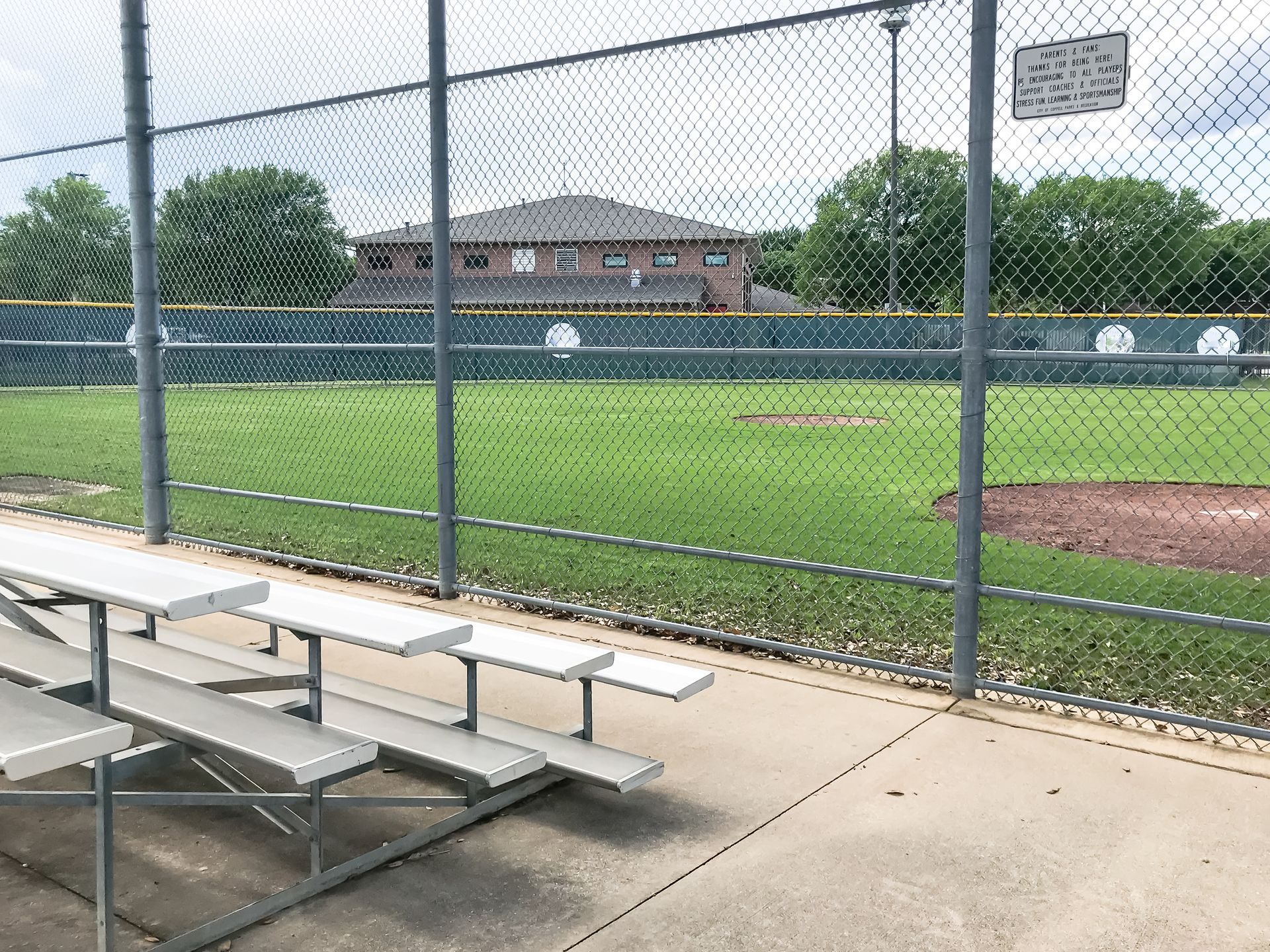 Empty metal bleachers sit on a concrete path in front of a chain-link fence bordering a grassy baseball field.