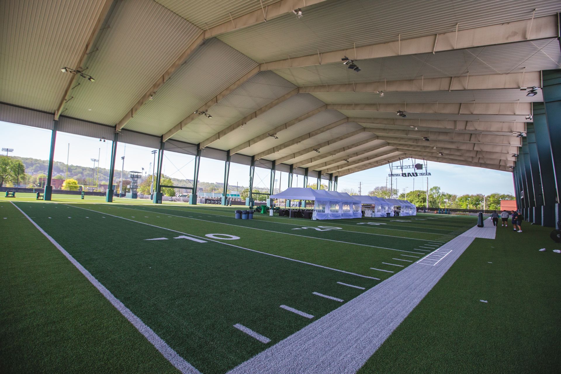 A wide-angle view of an indoor athletic field with green turf, white yard lines, and a large white tent on the sideline.