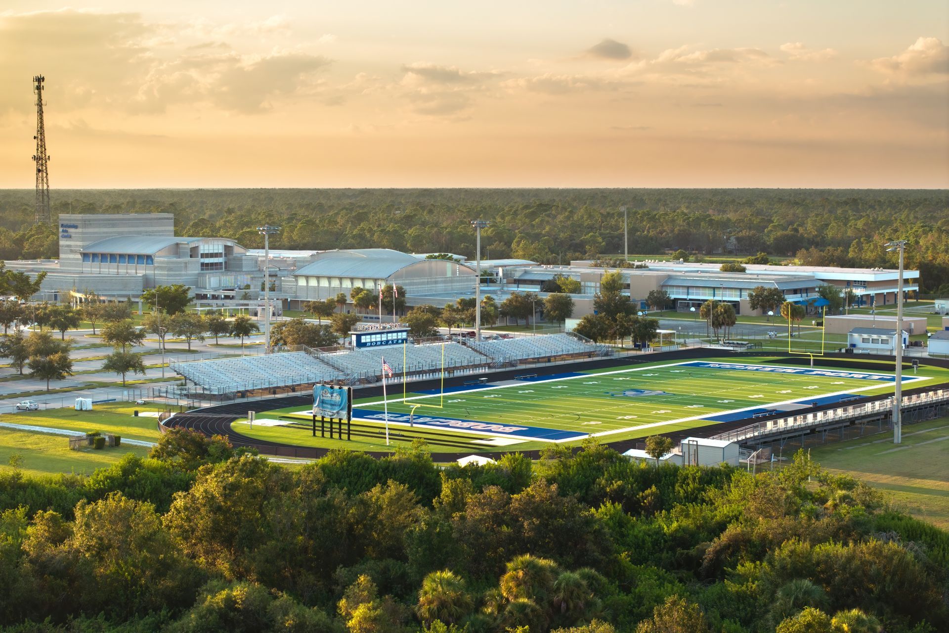 A high-angle view of a school campus with a football field in the foreground, surrounded by trees at sunset.