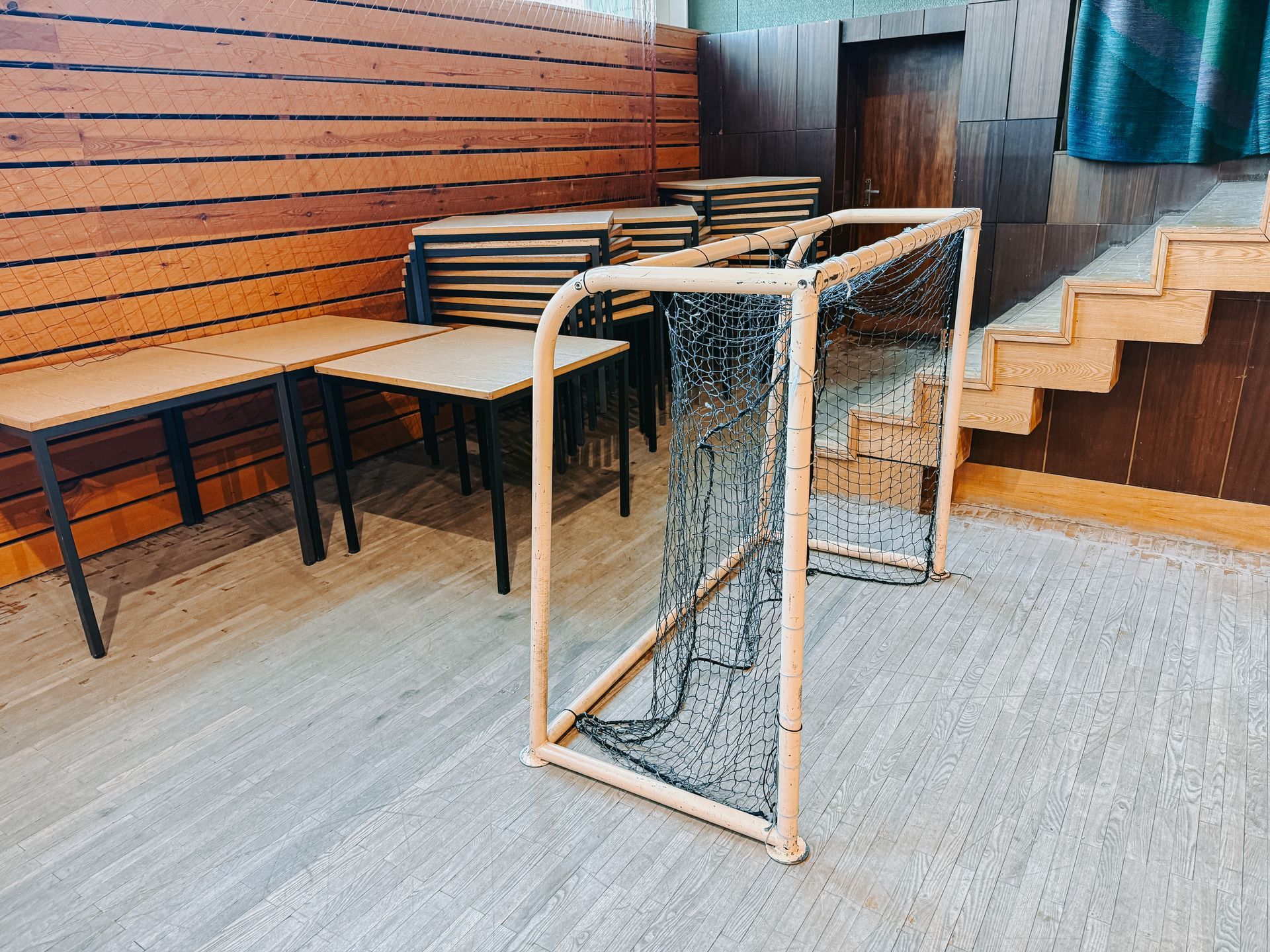 A small indoor hockey goal sitting on a floor next to a stack of wooden tables in front of a wood-paneled wall.