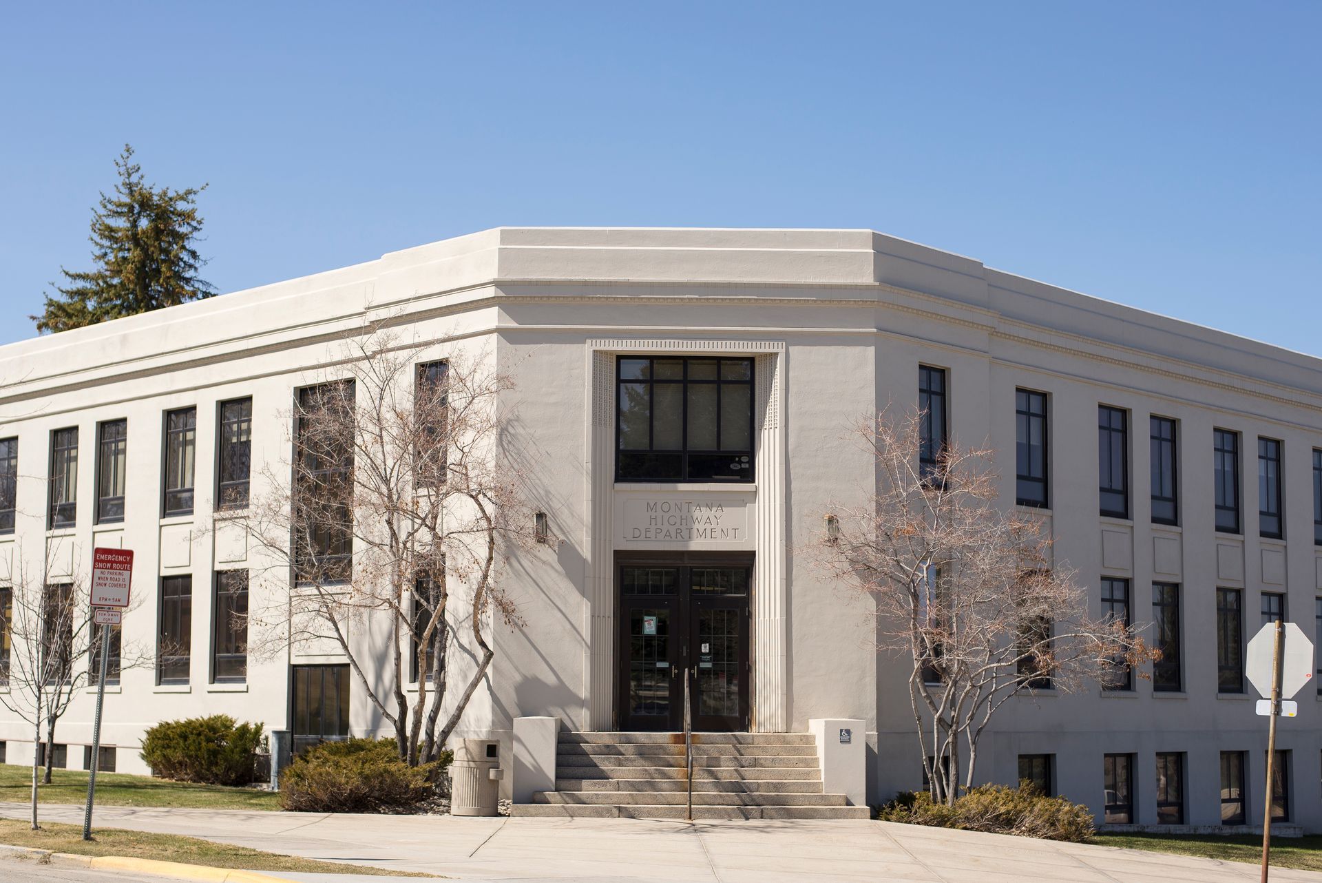 A two-story, white, L-shaped brick building with rectangular windows and a centered recessed entrance with stairs.