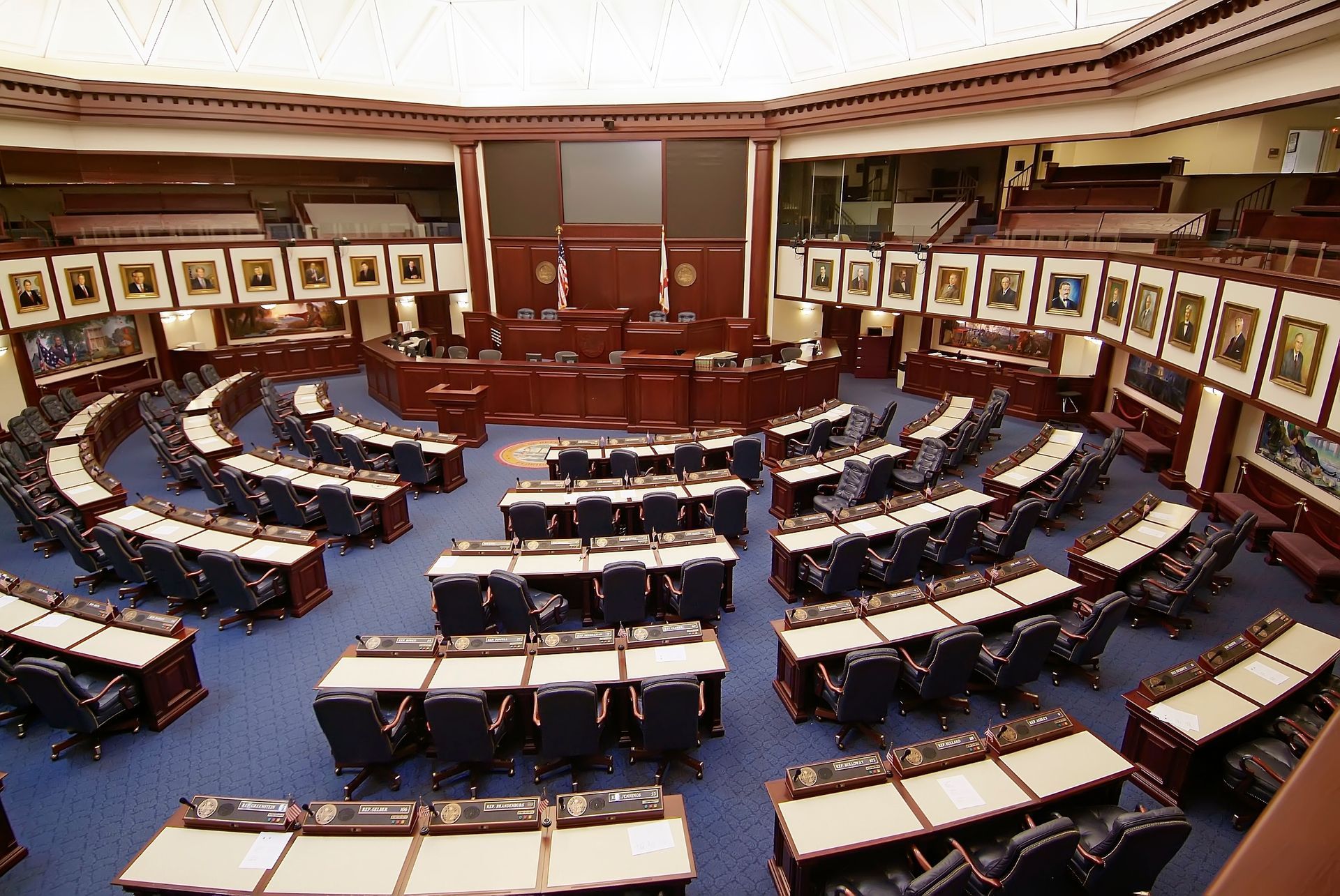 An empty government assembly hall featuring rows of wooden desks and blue chairs arranged in a semi-circle.