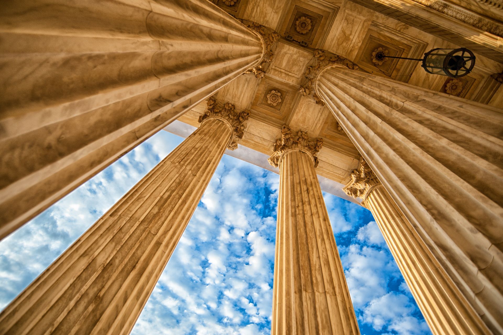 Low-angle view of grand stone columns leading up to an ornate ceiling against a bright blue sky with fluffy white clouds.