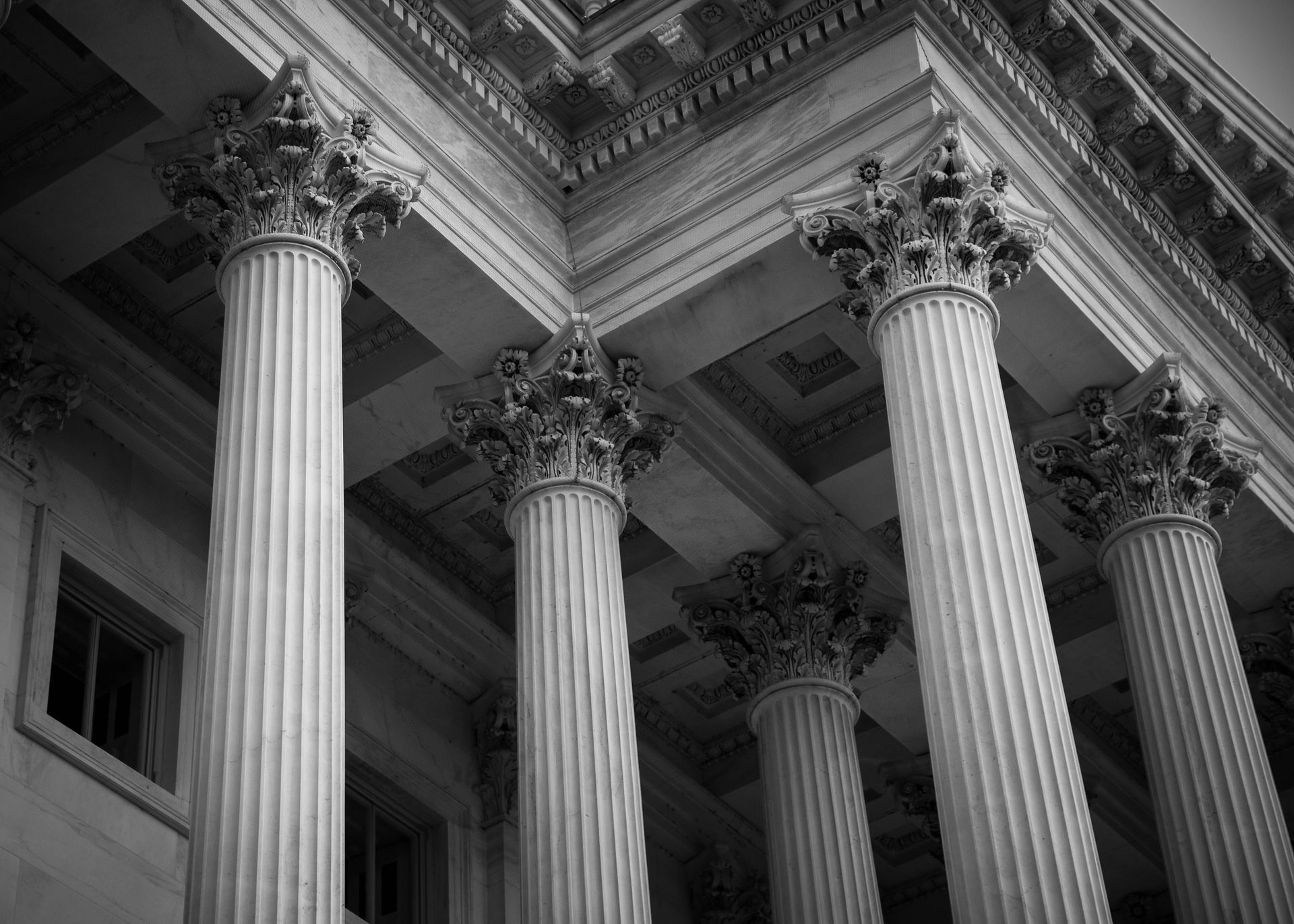 A low-angle, grayscale view of classical columns with ornate Corinthian capitals supporting a building's roof.