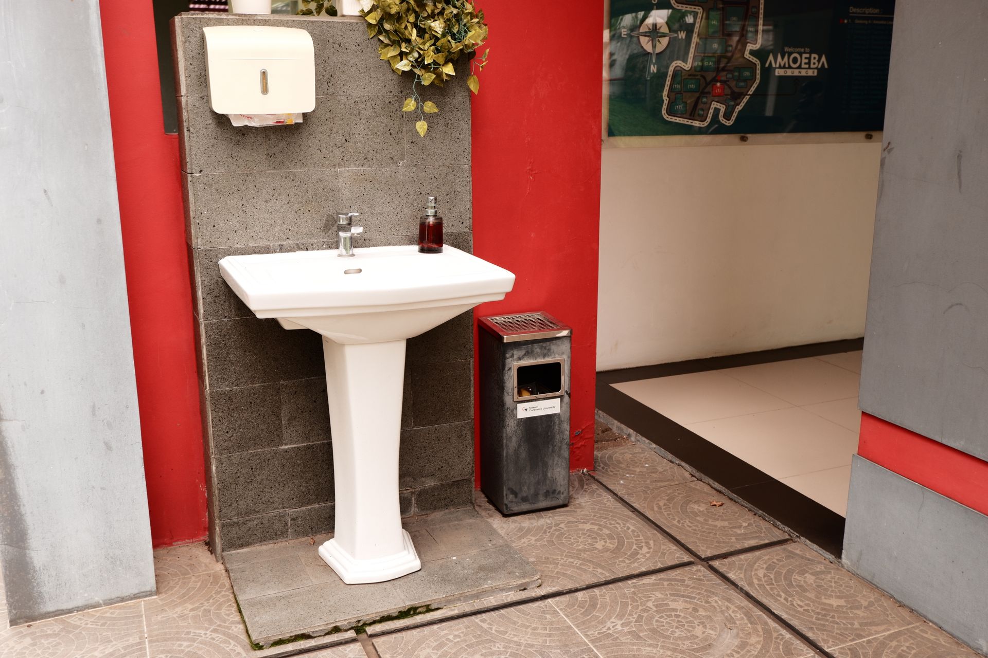 A white pedestal sink against a dark stone wall, beside a metal trash can and a red wall in a hallway.