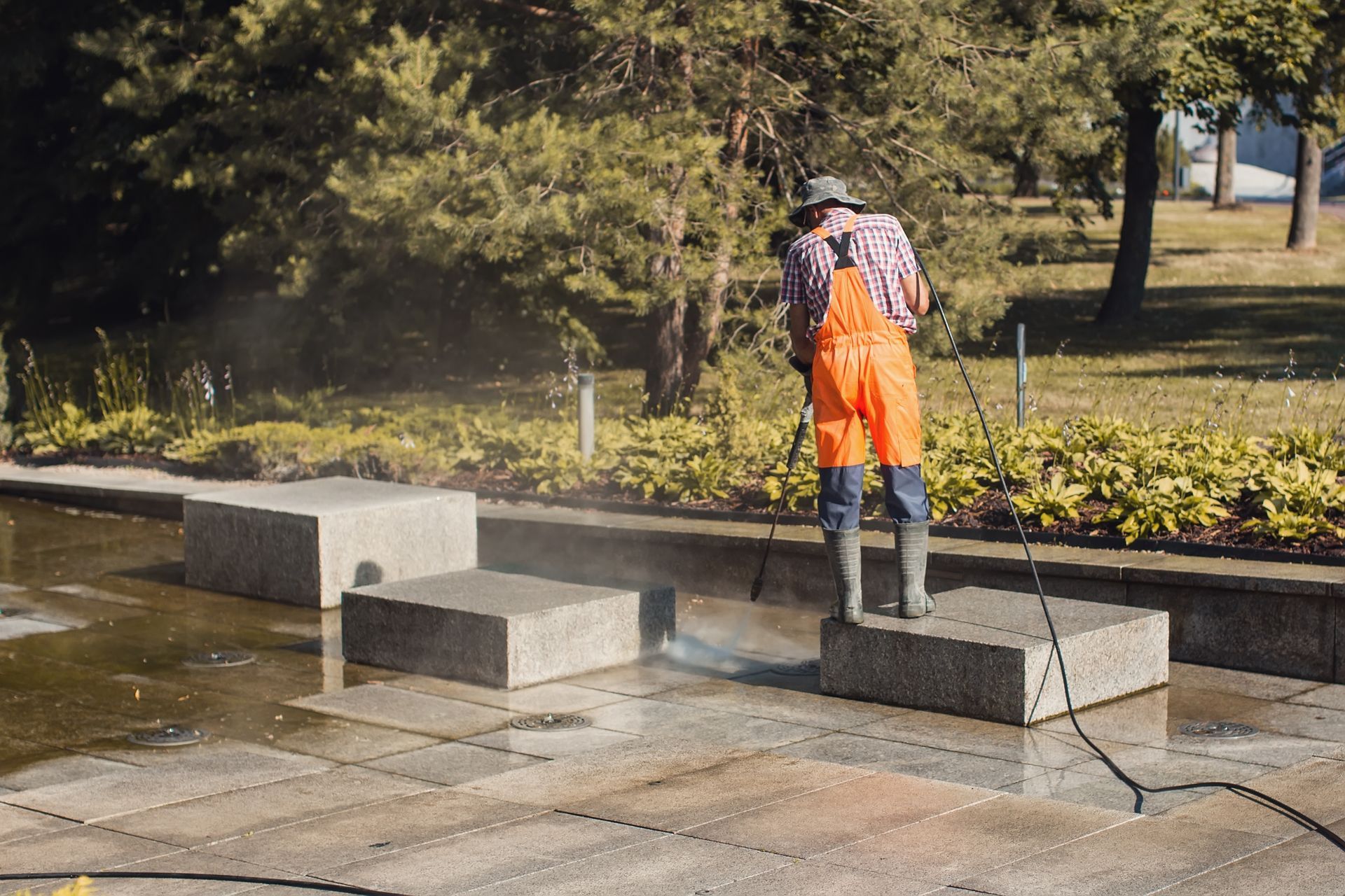 A person in bright orange overalls and boots uses a pressure washer to clean stone blocks in a public fountain area.