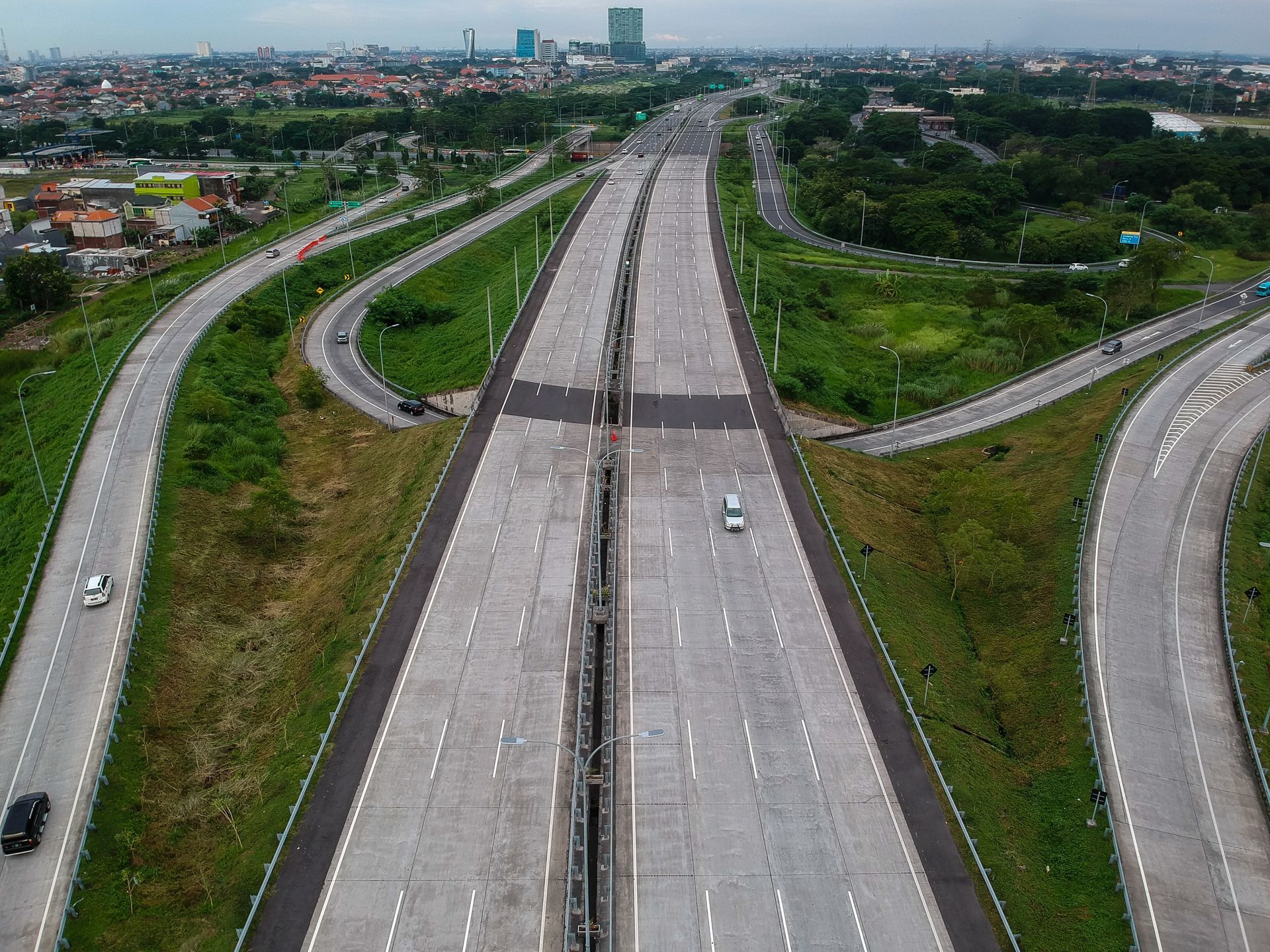 An aerial view of a multi-lane highway interchange with sparse traffic, surrounded by greenery and urban development.