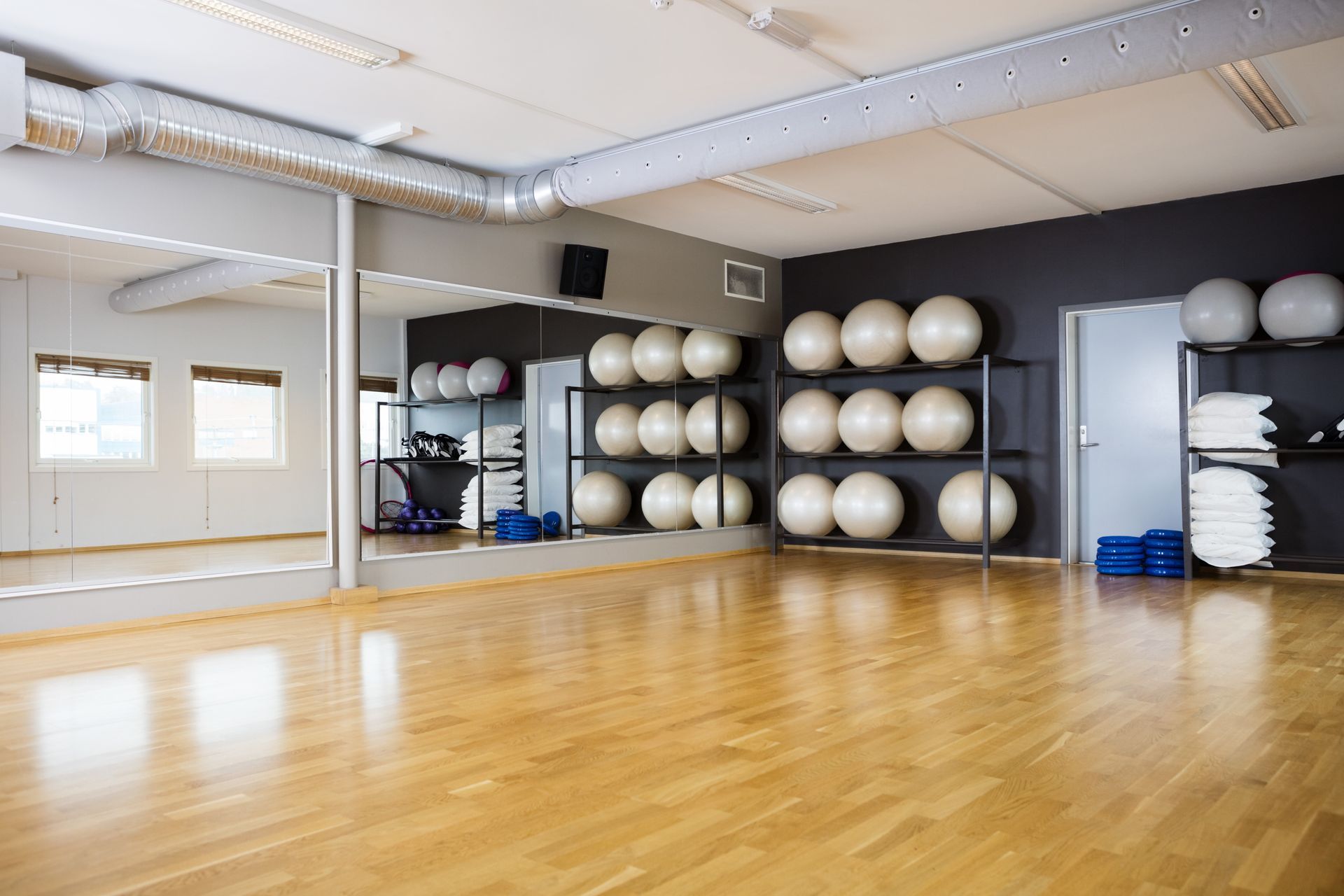 An empty gym studio with wood floors, wall mirrors, and shelving stocked with yoga and stability balls.