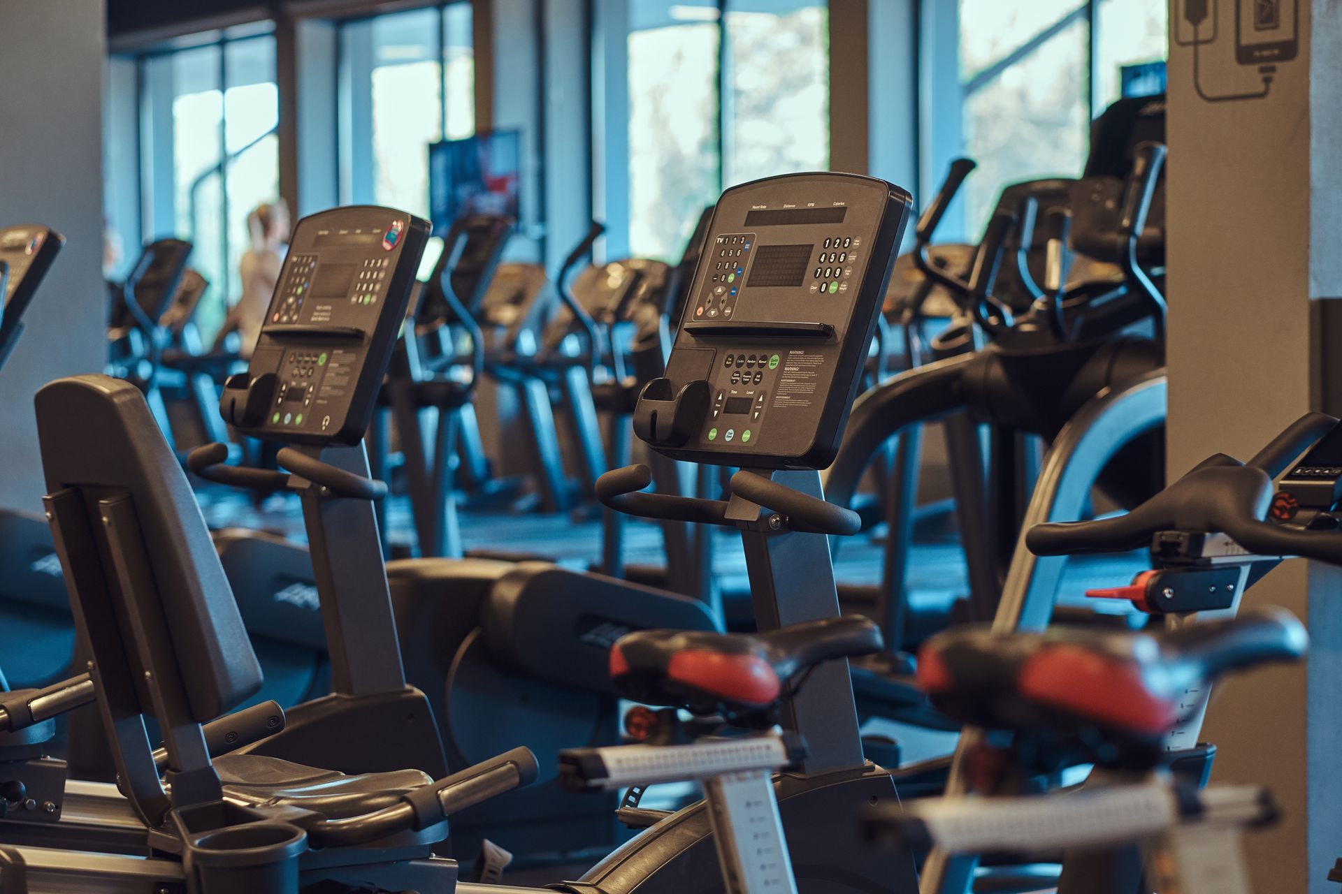 A row of stationary exercise bikes with digital consoles inside a gym, with more equipment visible in the background.