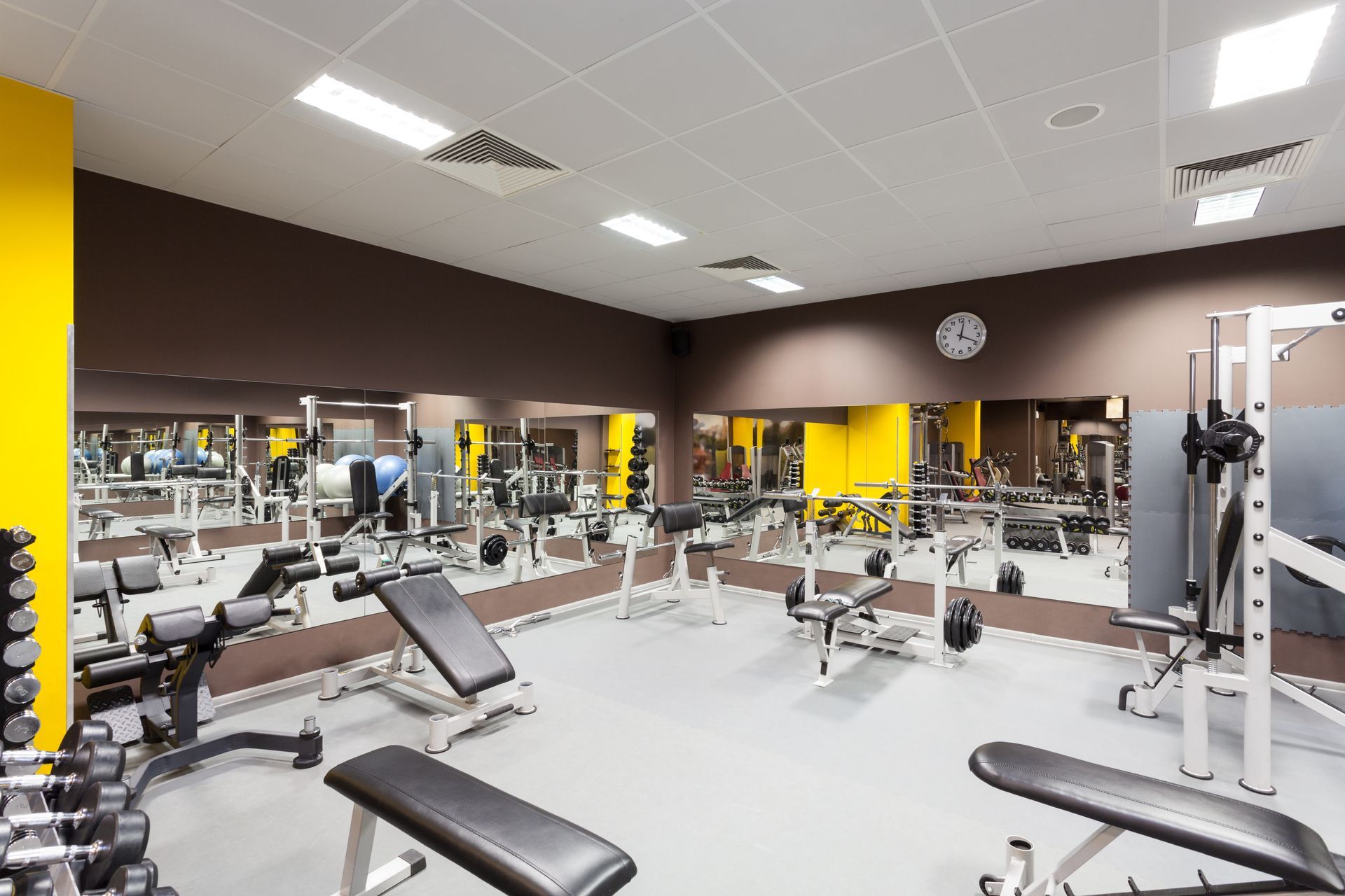 A brightly lit, modern gym with workout benches, mirrors, racks, and yellow accent walls.