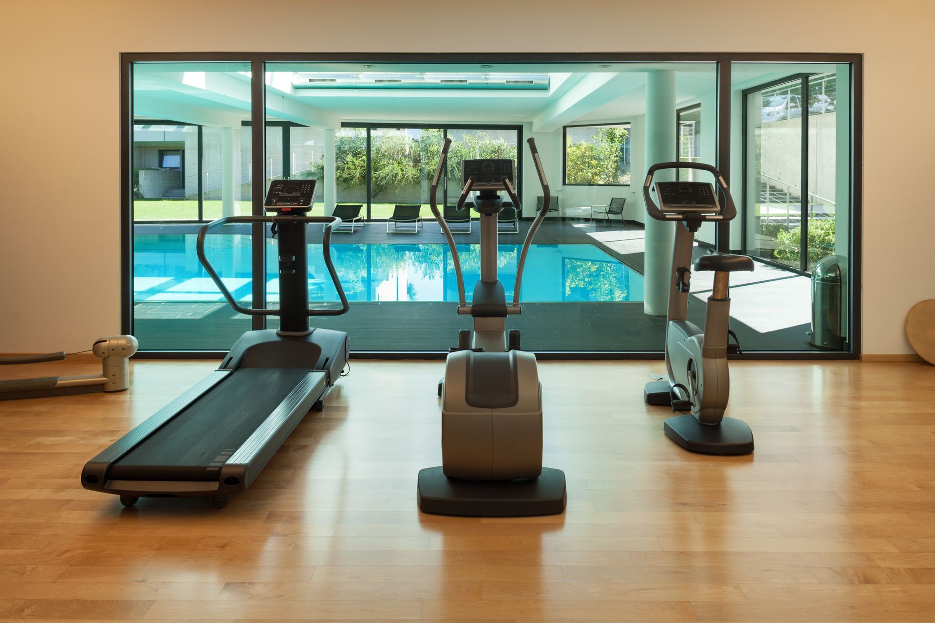 A gym with a treadmill, elliptical, and stationary bike, overlooking an indoor swimming pool through glass walls.