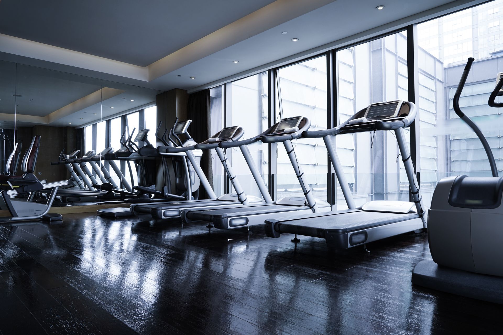 A row of treadmills facing floor-to-ceiling windows in a modern, dark-floored gym.