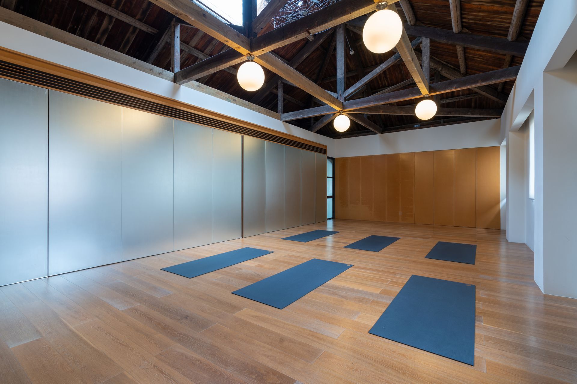 A brightly lit yoga studio with wooden floors, an exposed beam ceiling, and six blue mats arranged for a class.
