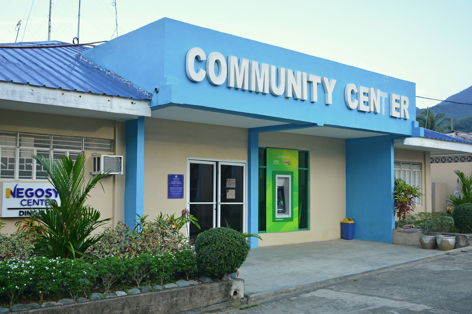 A blue and tan community center building with an outdoor ATM and a small garden area.